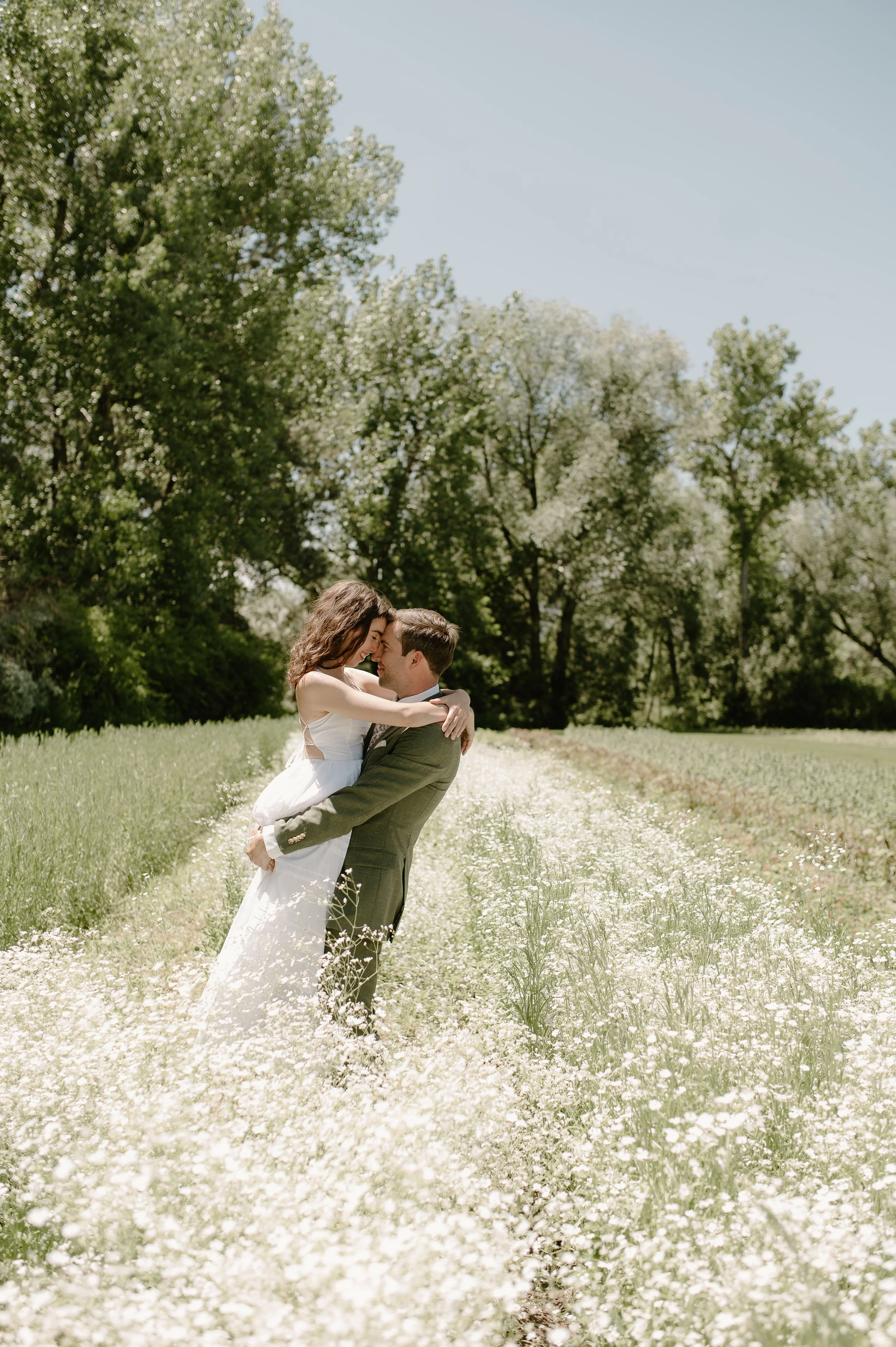 A couple in wedding attire embracing in a field of white flowers, with green trees and a clear blue sky in the background.