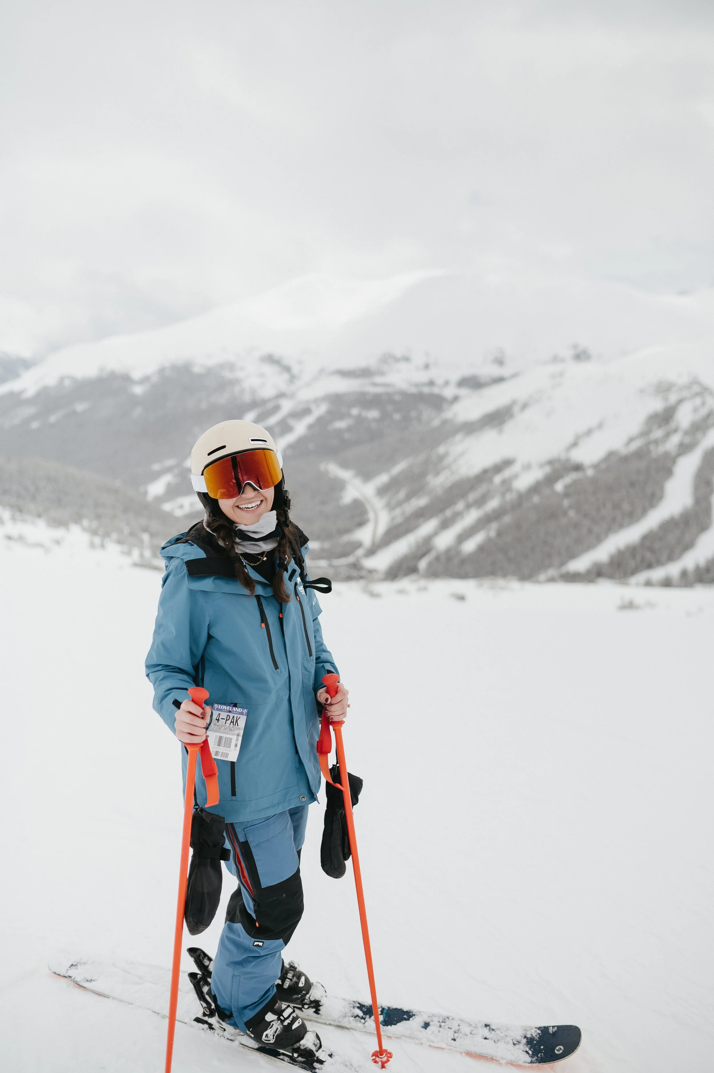 A person dressed in blue ski gear holding ski poles, smiling while standing on snow with a mountainous, snow-covered landscape in the background.