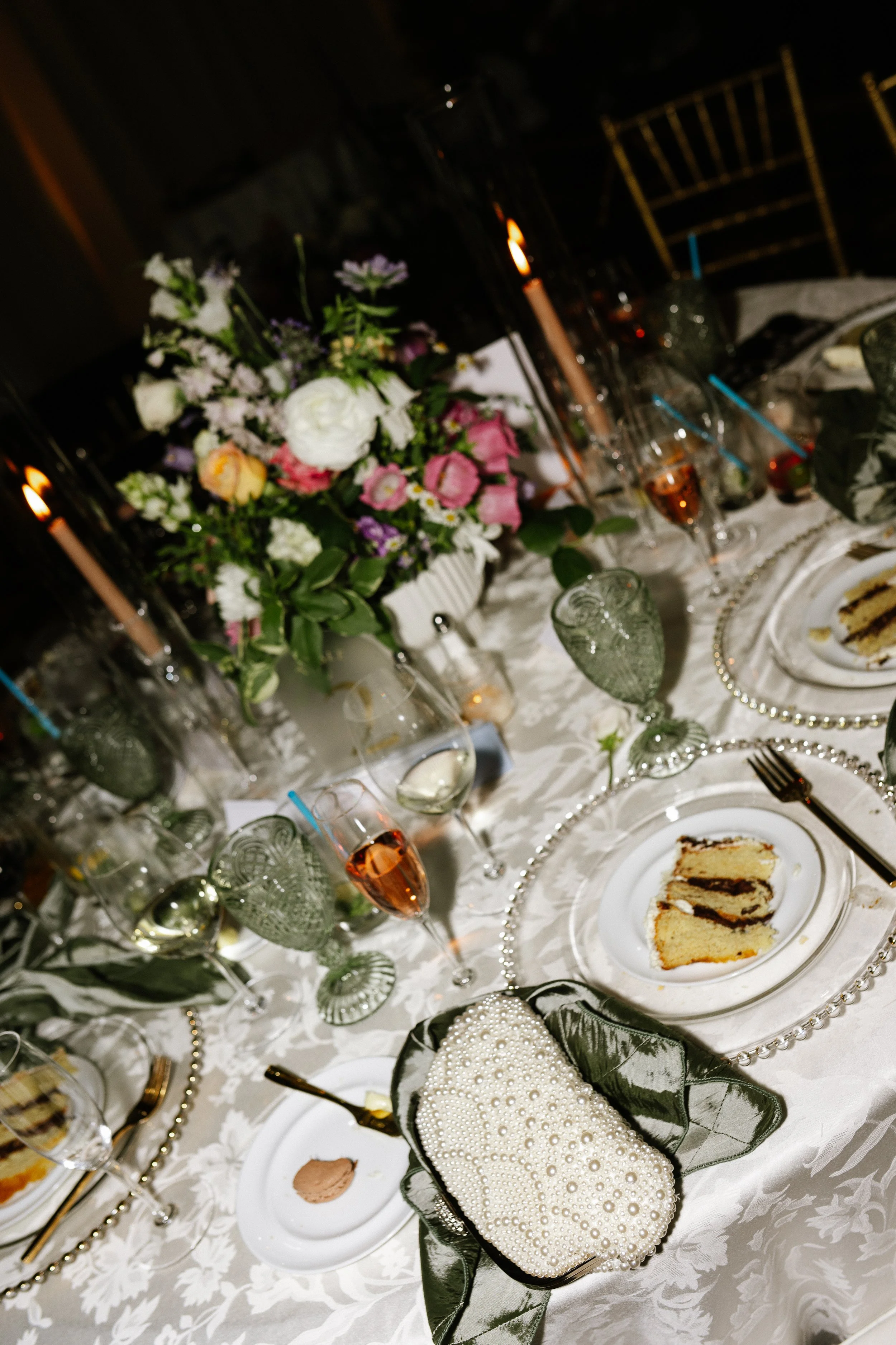 Elegant table setting with floral centerpiece, candles, champagne glasses, plates with cake, and decorative pearl clutch in a formal dining setting.
