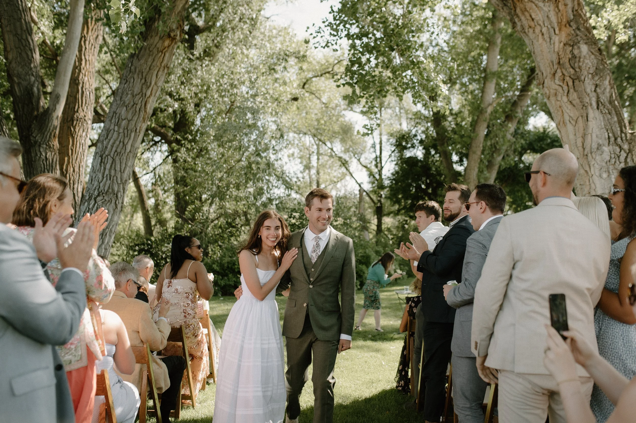 bride and groom walking down an aisle under trees surrounded by their guests clapping