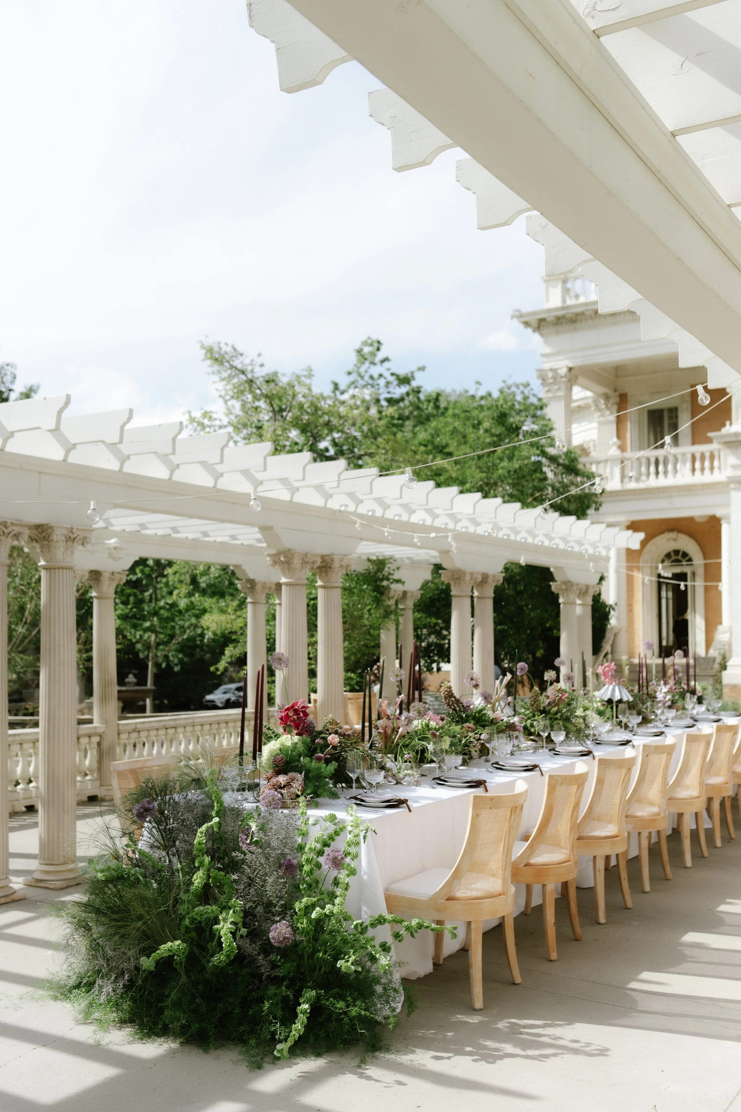 photo of a wedding reception set up outside of a mansion in Denver, Colorado. Image features a long rectangular table set with florals, candles, and a china set