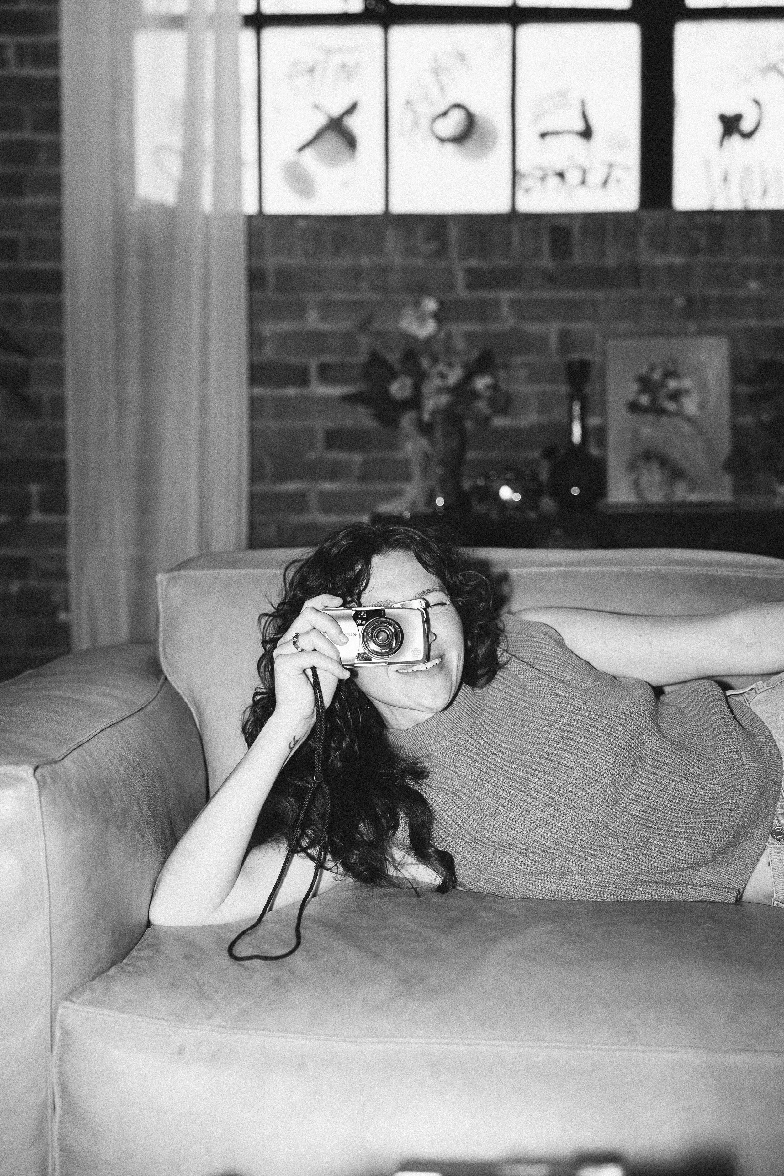A woman with long curly hair lying on a couch, holding a camera to her eye as if taking a photo, with a brick wall, framed pictures, a window, and a bookshelf in the background.