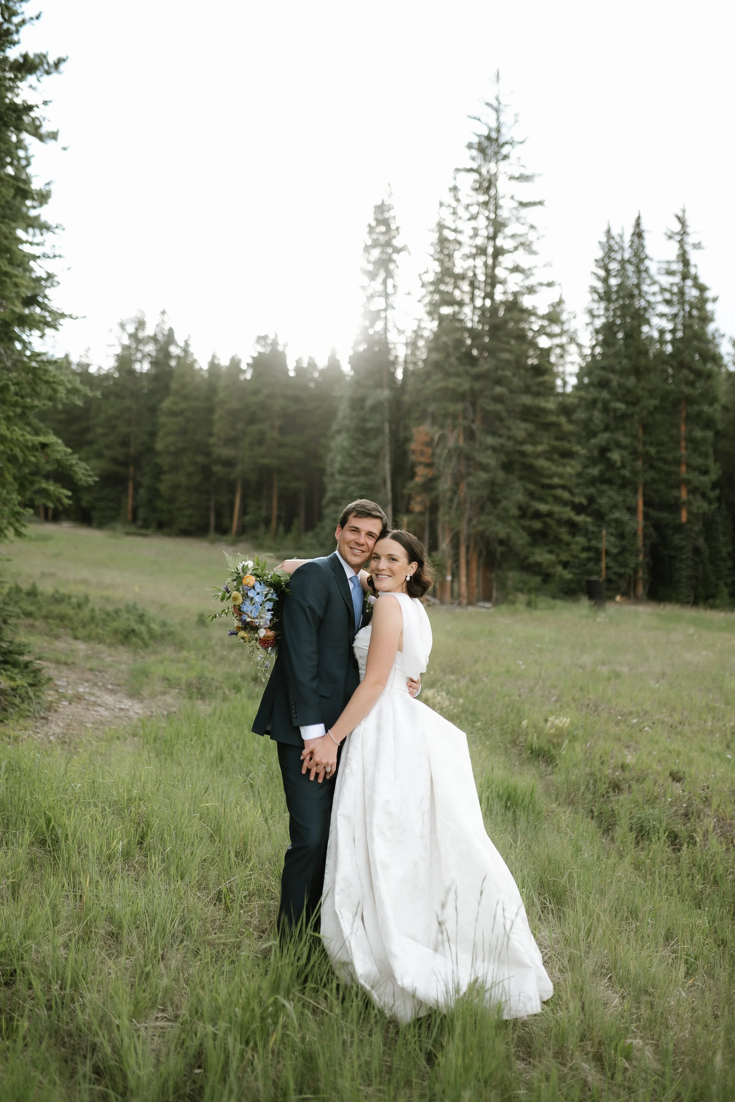 A bride and groom standing together in a grassy field with tall trees in the background, smiling and holding hands, outdoors on a bright day.