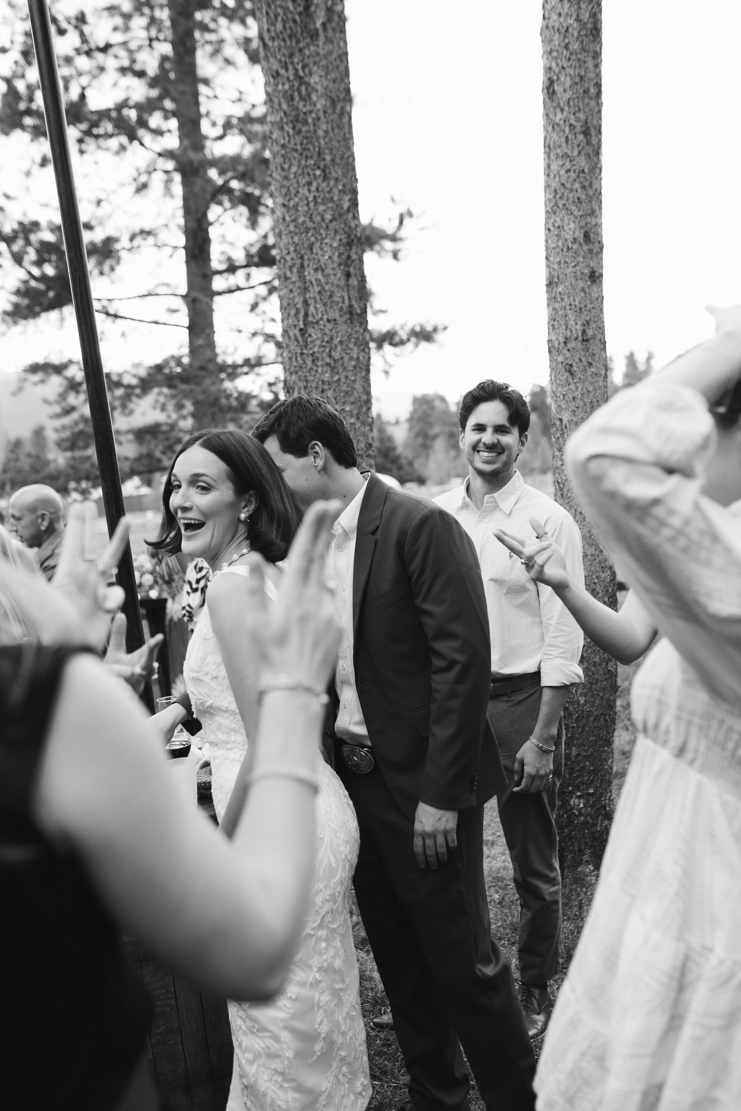 Black and white photo of a woman in a wedding dress smiling and looking at camera, with a man in a suit and others in the background outdoors near trees, at a wedding reception or similar event.