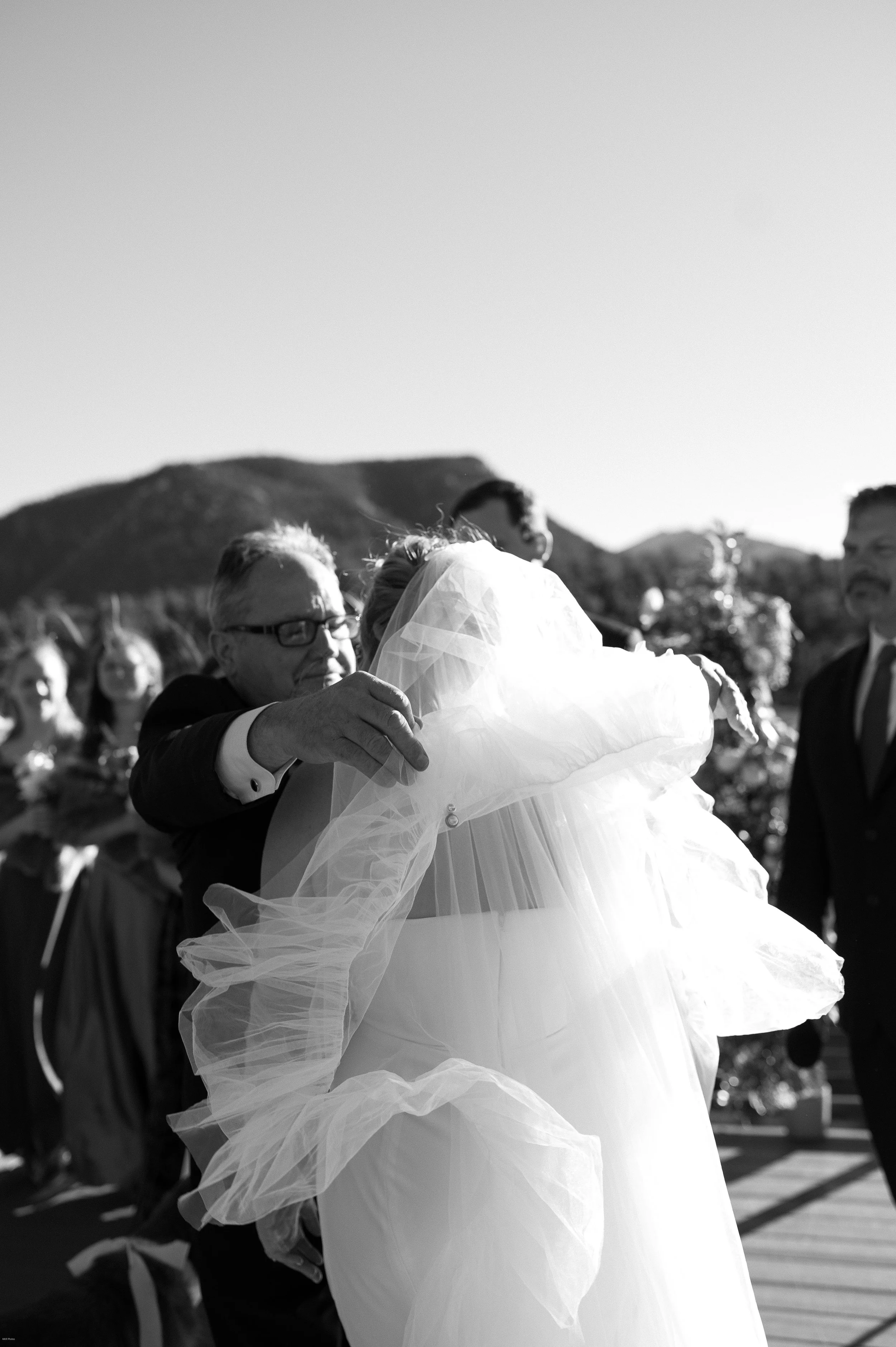 A wedding scene with a bride in a white dress and veil hugging an older man in a suit. Several guests are in the background outdoors with mountains and clear sky.