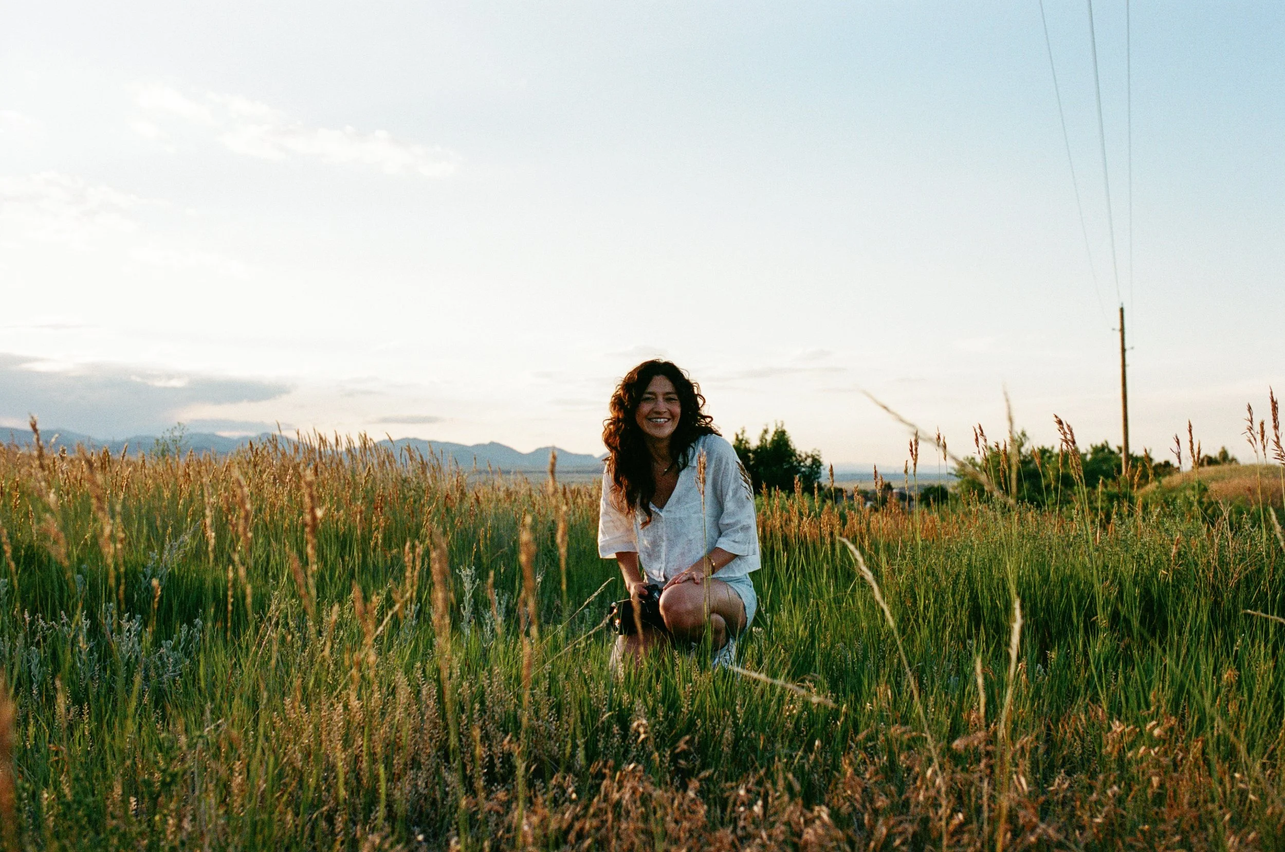 A woman with curly dark hair, smiling, crouching in a field of tall grass during sunset, with mountains and a power line in the background.