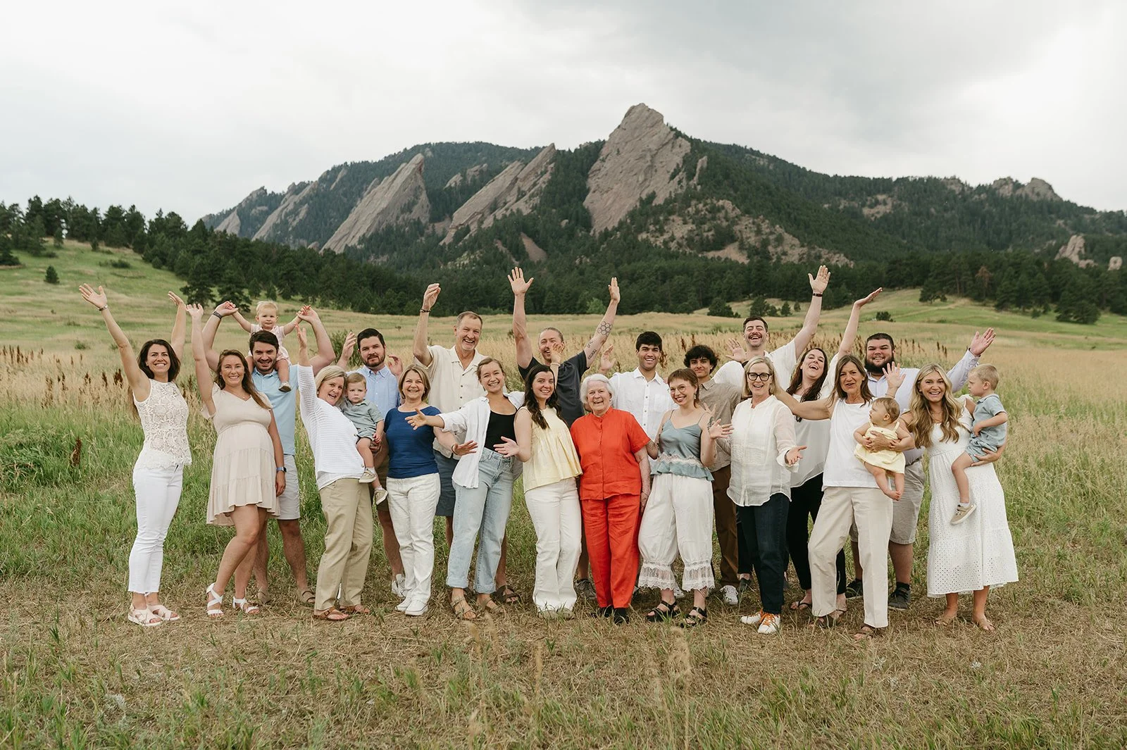 A large group of people, including children, gathered outdoors in a field with mountains in the background. They are smiling, with some raising their hands, celebrating, or holding children.