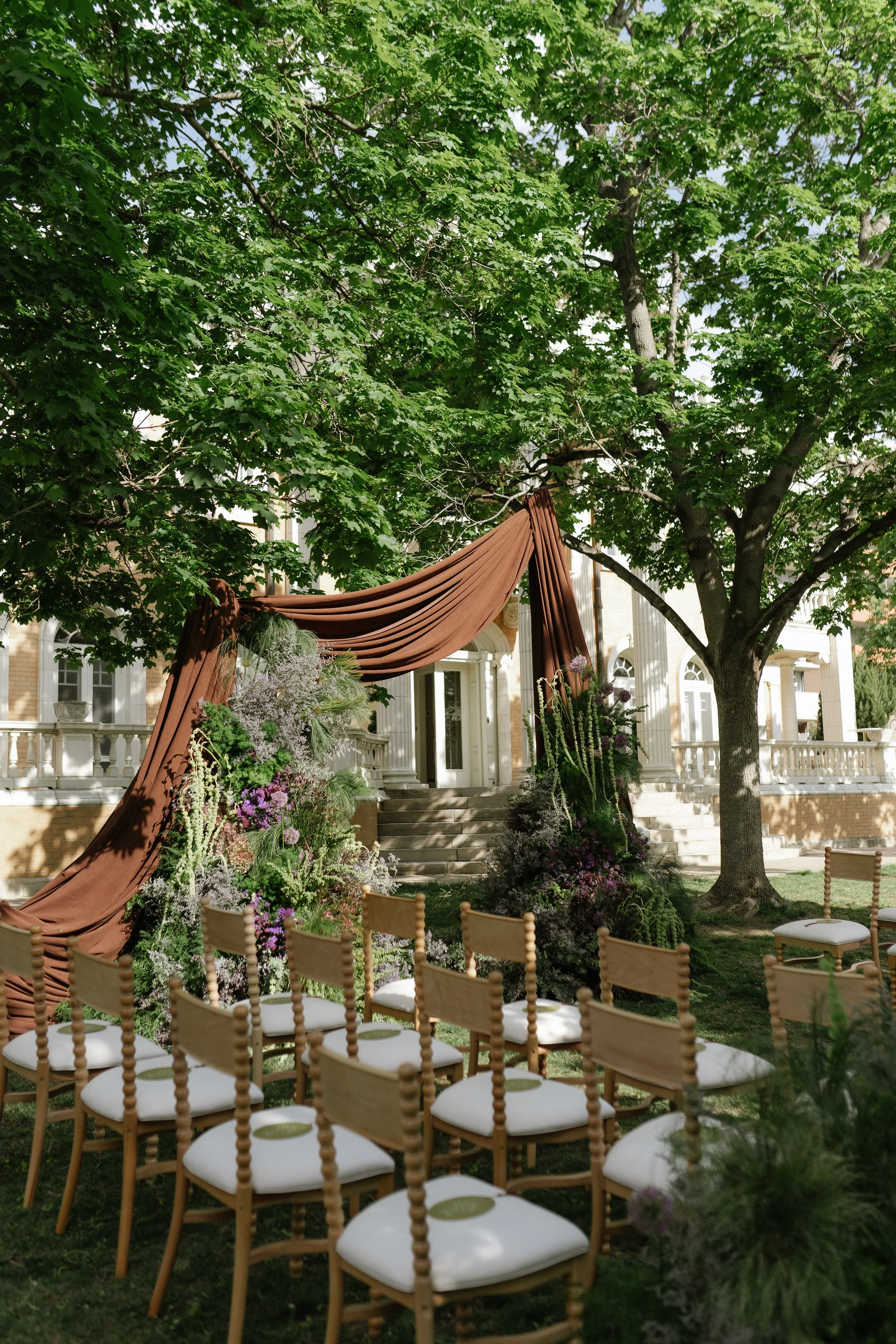 photo of a wedding ceremony set up in front of a house with pillars and under the shade of trees. There are rows of wooden chairs facing a wedding arch of drapery and green and purple flowers