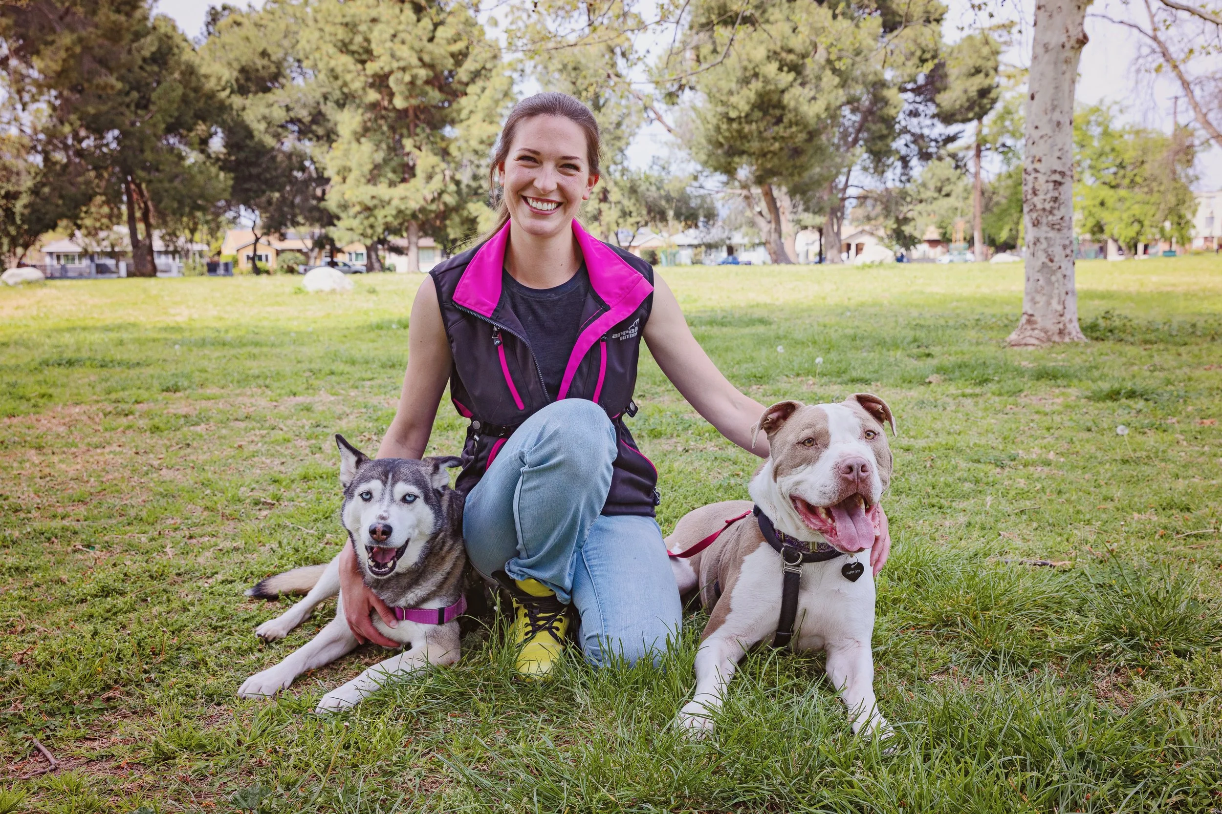 Vic kneeling in the grass with a husky (Lovely) and a pit bull (Dougal) laying beside her