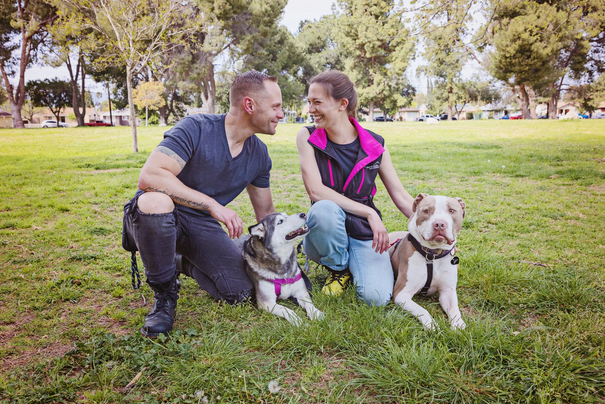 Vic and partner kneeling in the grass with two dogs, a husky and a pit bull, laying next to them