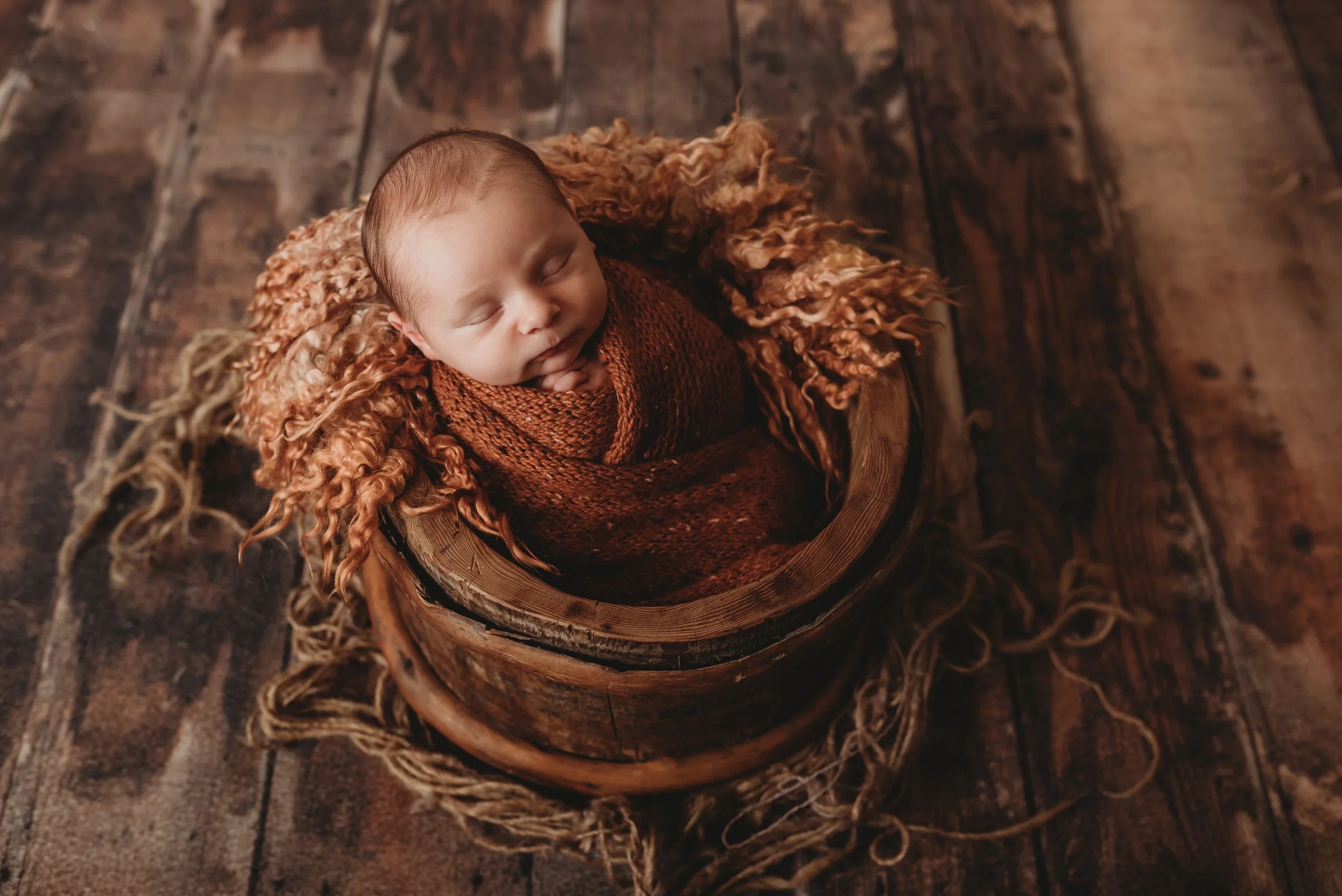 Sleeping newborn wrapped in brown cloth in wooden basket on rustic wooden floor.