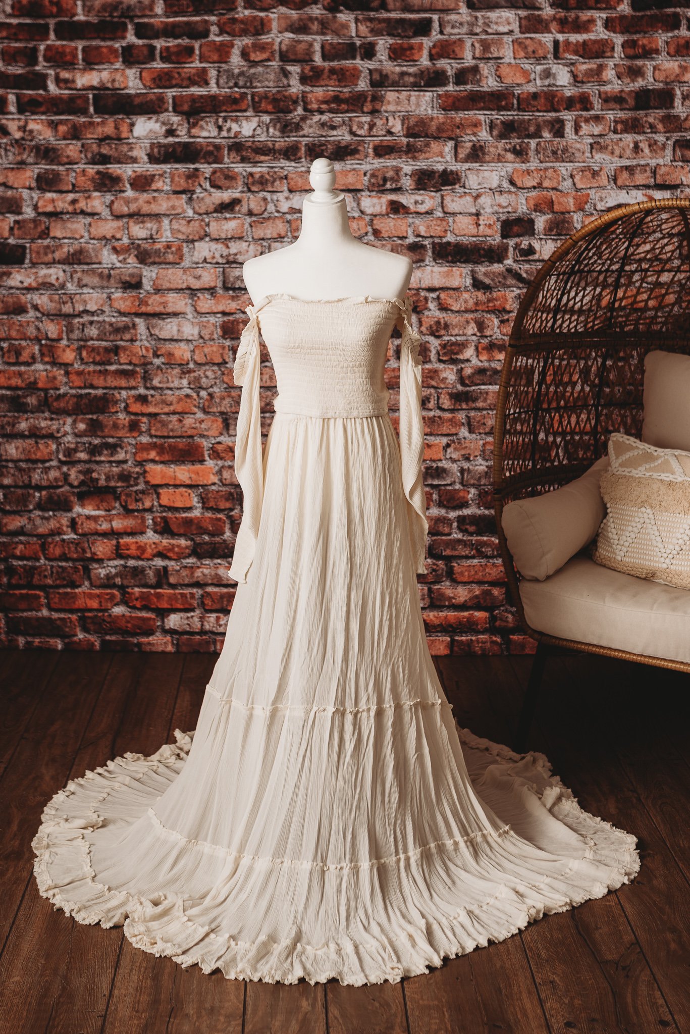 White wedding dress displayed on a mannequin against brick wall backdrop and wooden floor, with rattan chair visible.