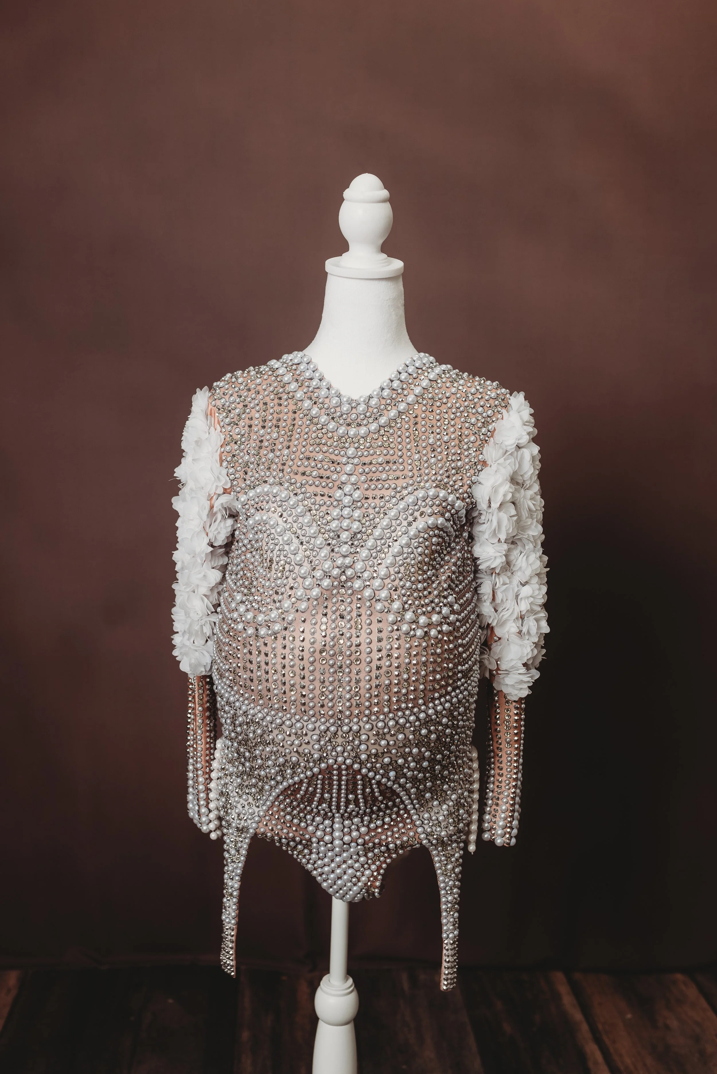 A mannequin dressed in a form-fitting dress decorated with numerous pearls, sequins, and white floral accents on the sleeves. The dress has intricate beading and pearl designs, and the mannequin stands on a white pedestal against a plain brown backgr