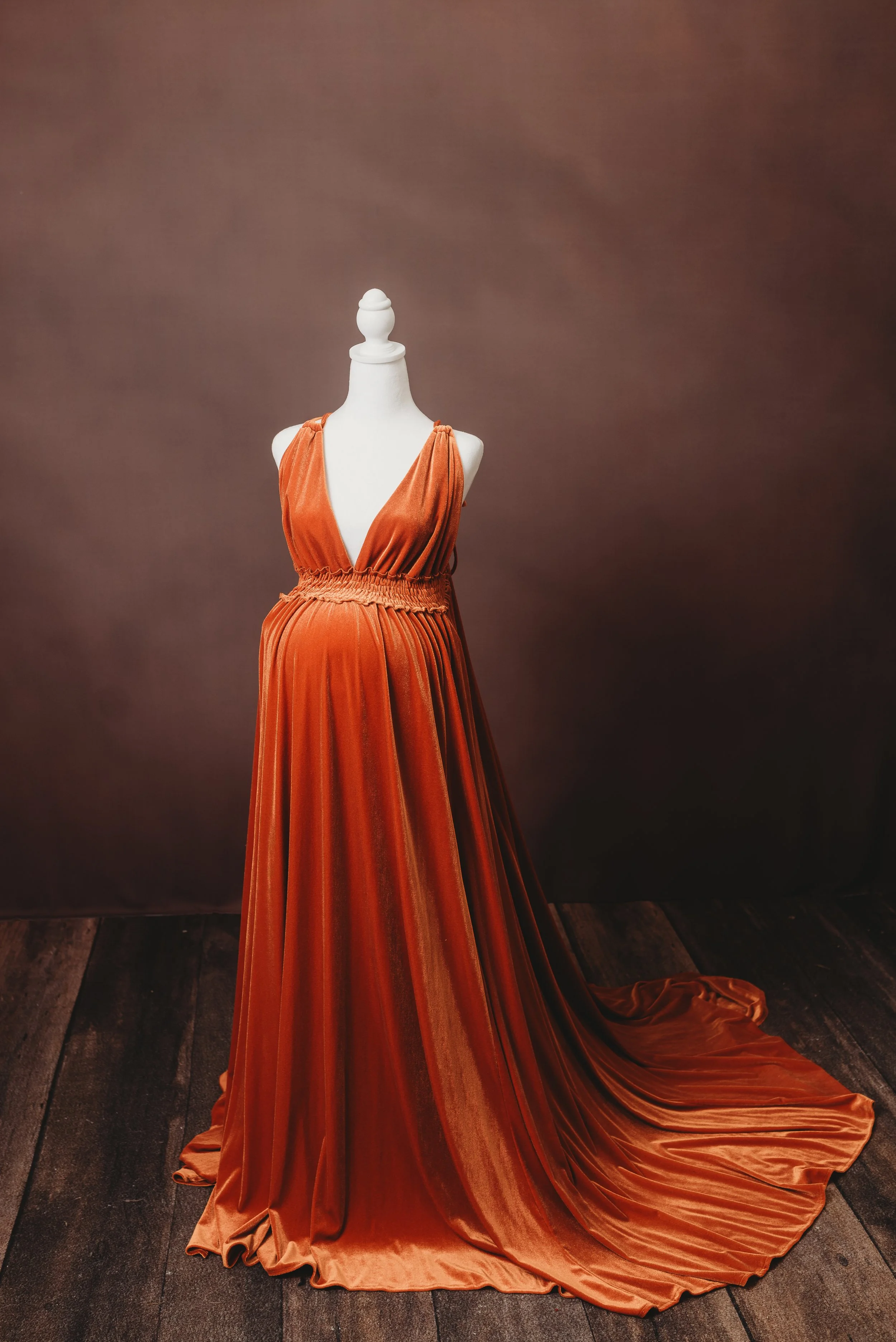 A mannequin dressed in an elegant, flowing orange velvet evening gown with a deep V-neckline, gathered waist, and a long train on a wooden floor against a dark brown background.