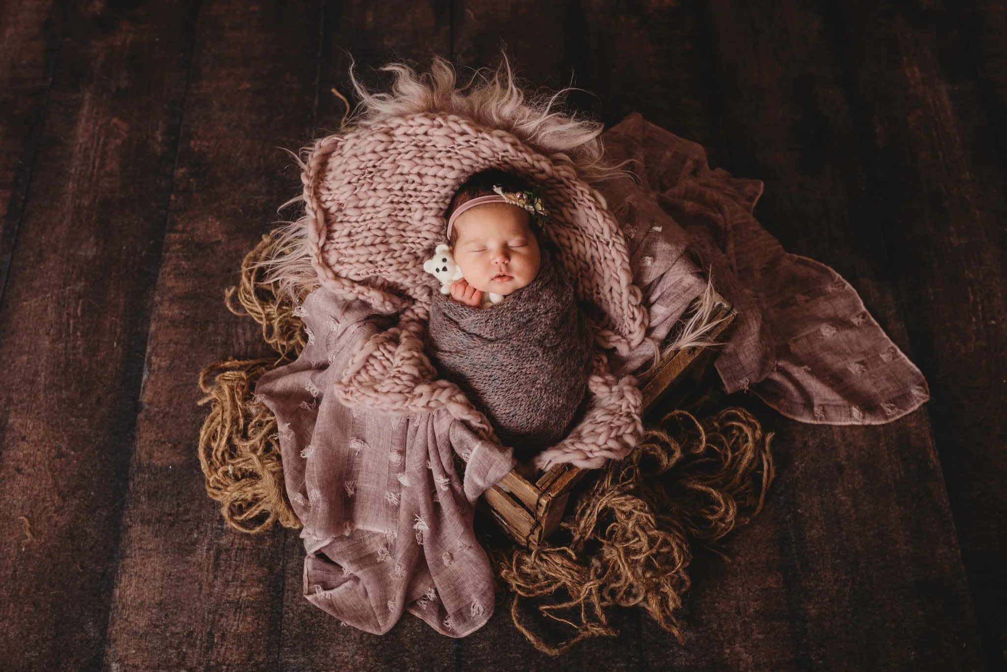 Newborn baby wrapped in a blanket, lying on a textured surface, with a stuffed toy, surrounded by soft fabrics.