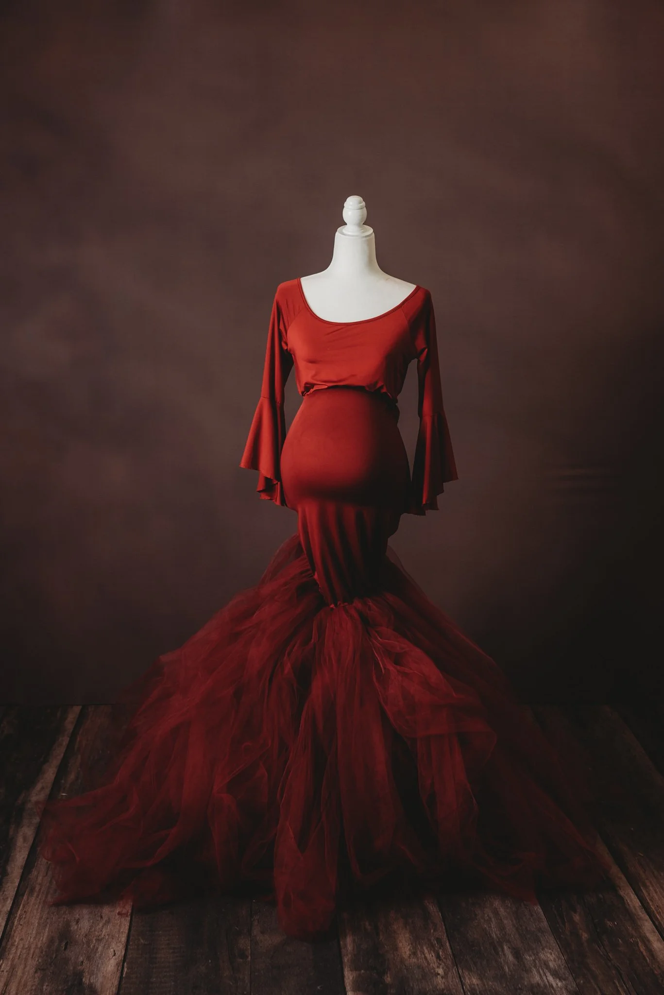 Red mermaid-style gown on mannequin with flared sleeves and tulle bottom; rustic wooden floor and dark background.