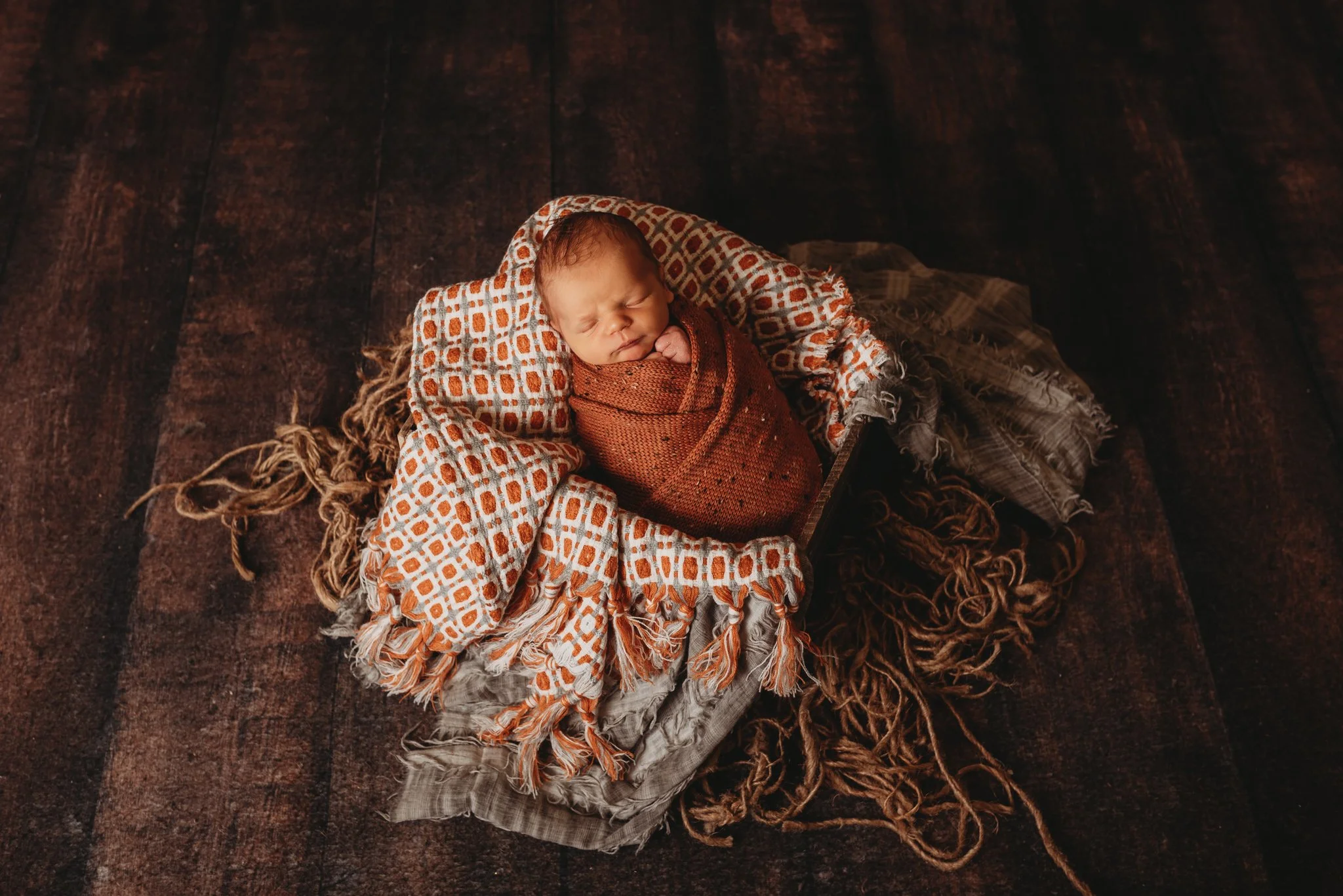 Newborn baby swaddled in a textured blanket, asleep on a rustic wooden surface with decorative fabric and twine.