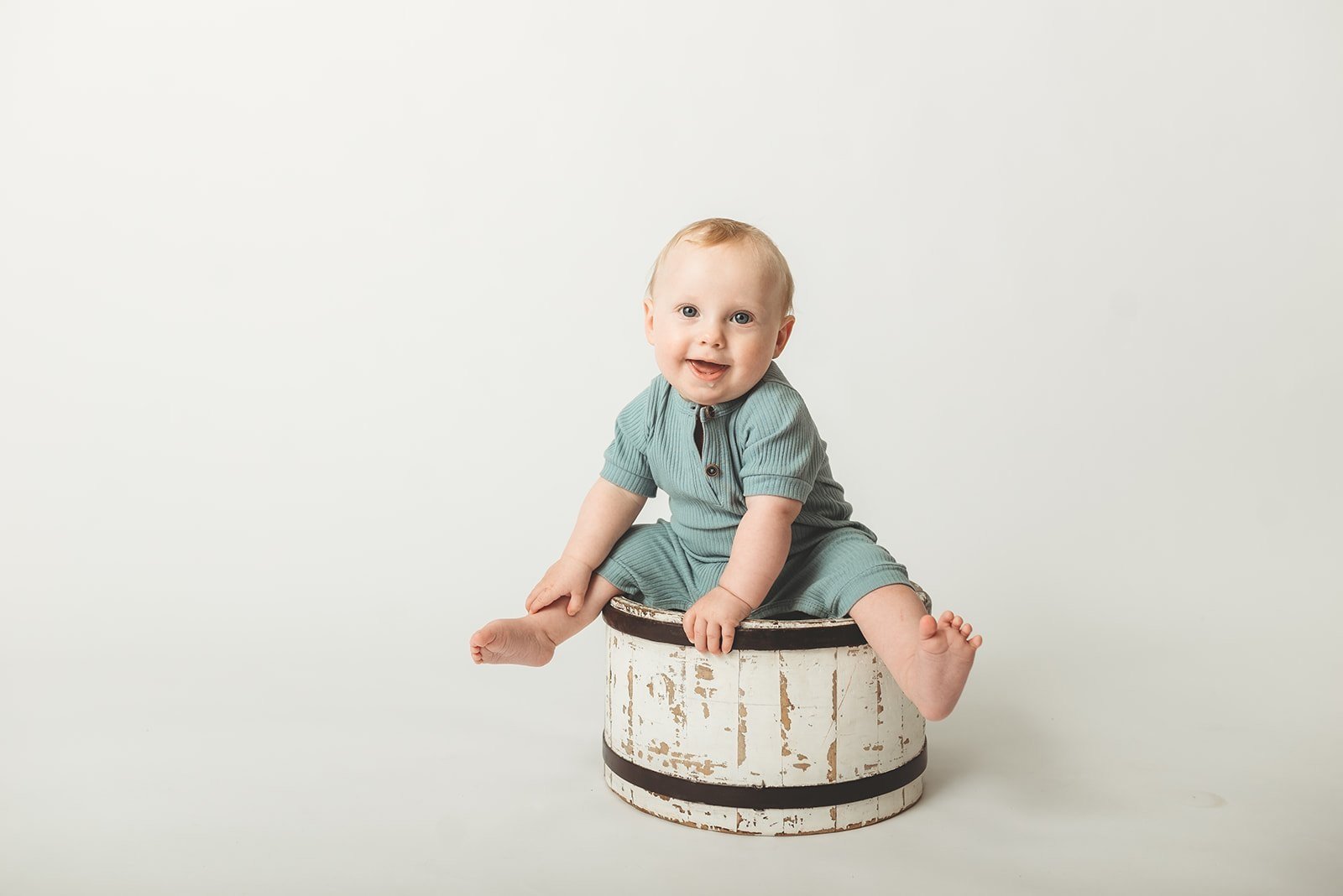Smiling baby sitting on a rustic wooden barrel, wearing a blue romper, against a light background.