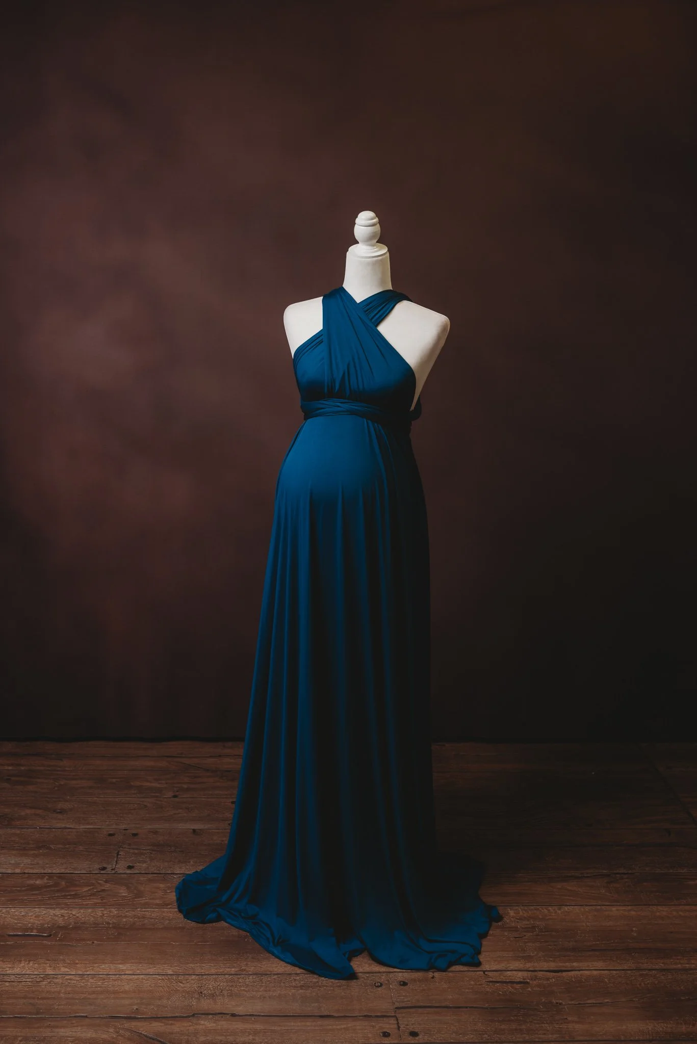 Elegant dark blue maternity dress on mannequin with wooden floor and brown background.