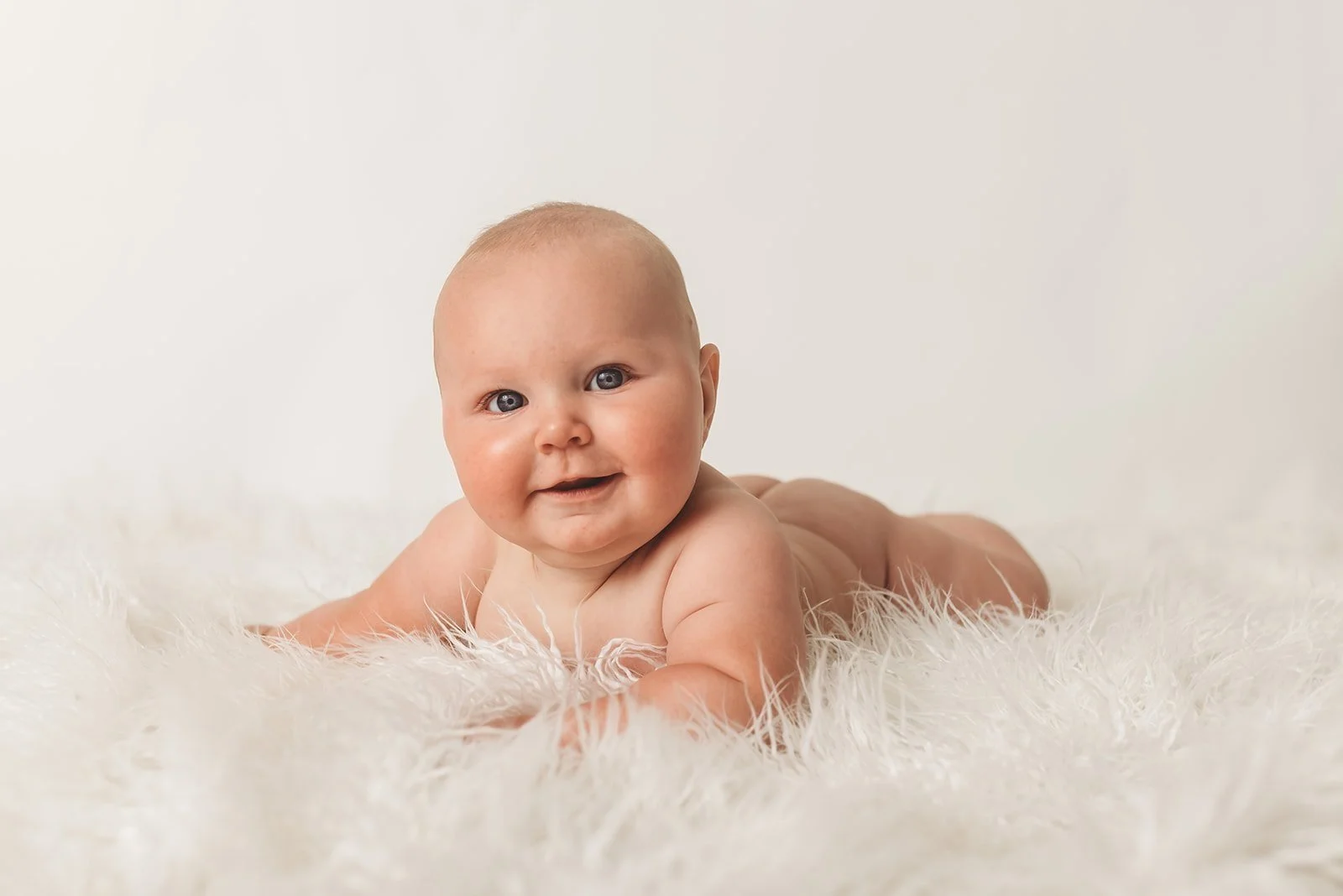Smiling baby lying on a fluffy white rug, facing the camera.