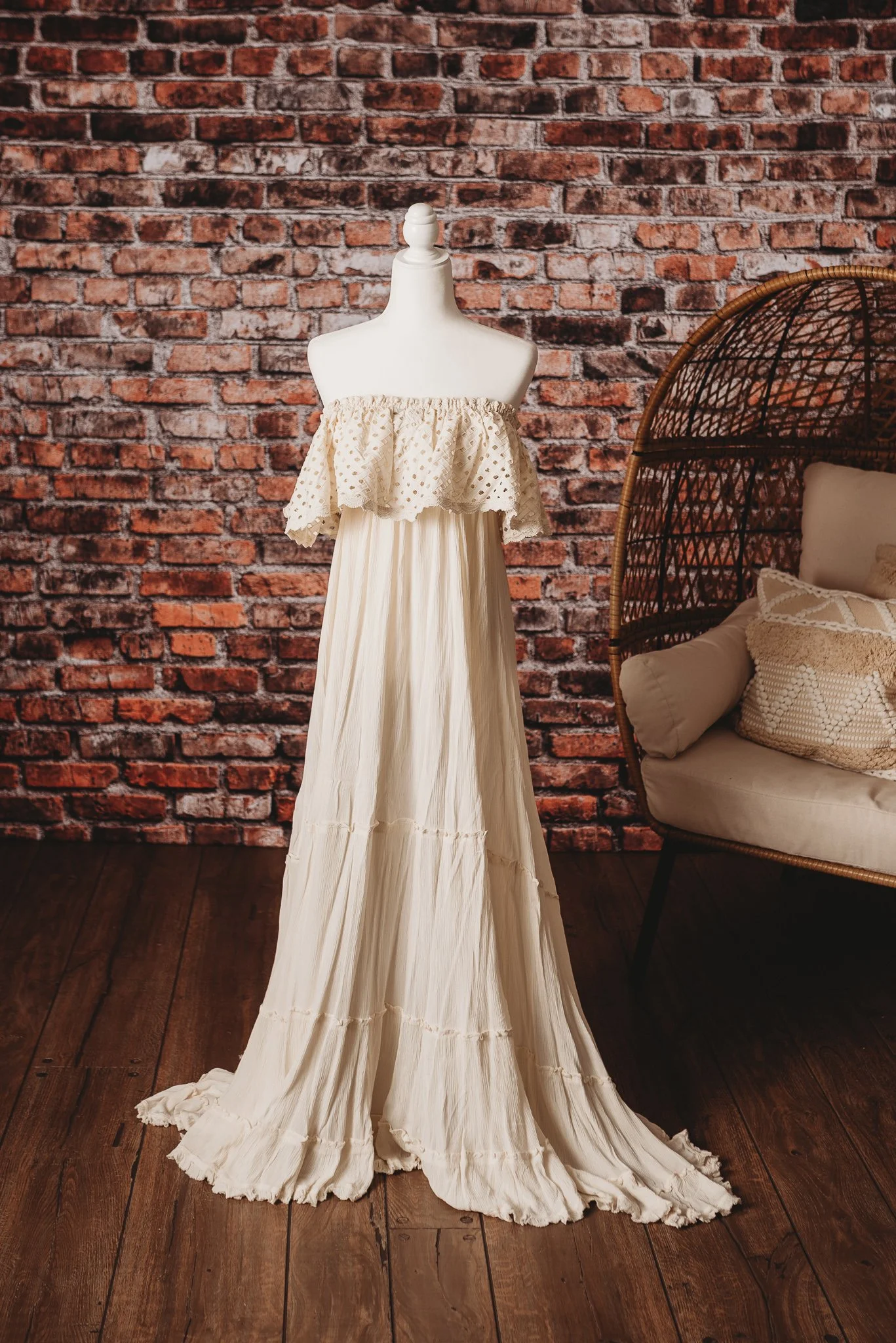 Off-shoulder beige maxi dress on mannequin with brick wall background, wicker chair, and cushions.