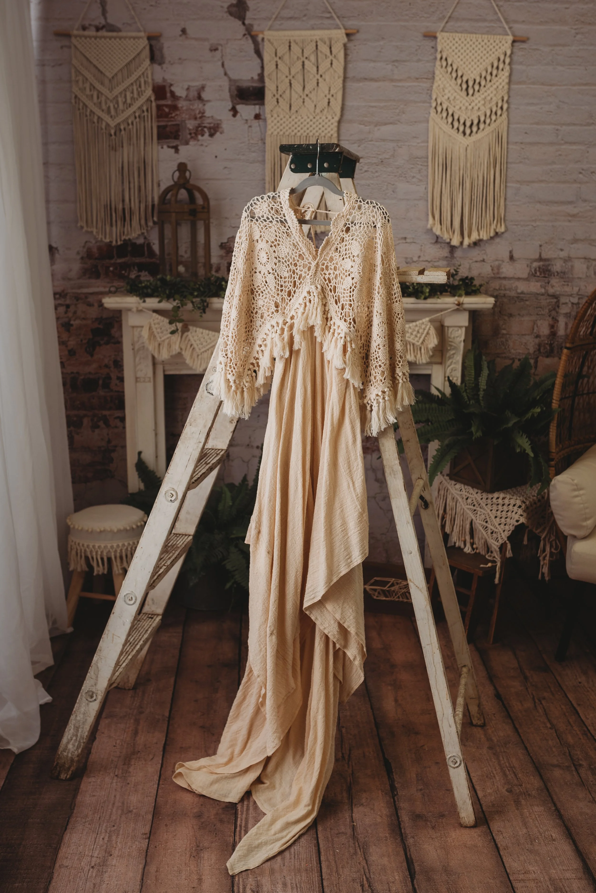 Bohemian style living room with a wooden ladder displaying a lace shawl and flowing beige fabric, surrounded by macrame wall hangings and potted plants.
