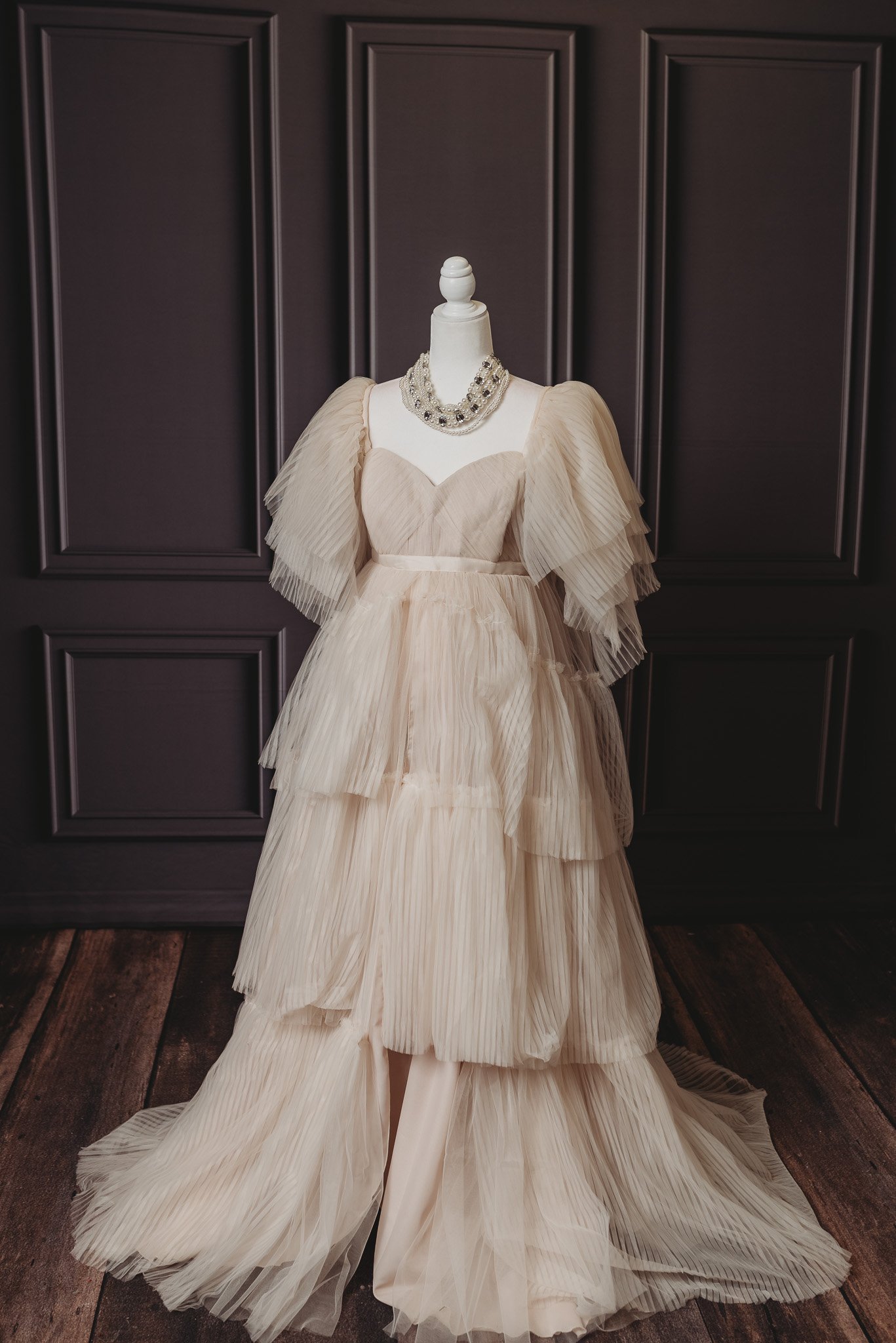Elegant beige tiered gown on mannequin with beaded necklace, dark panel backdrop.
