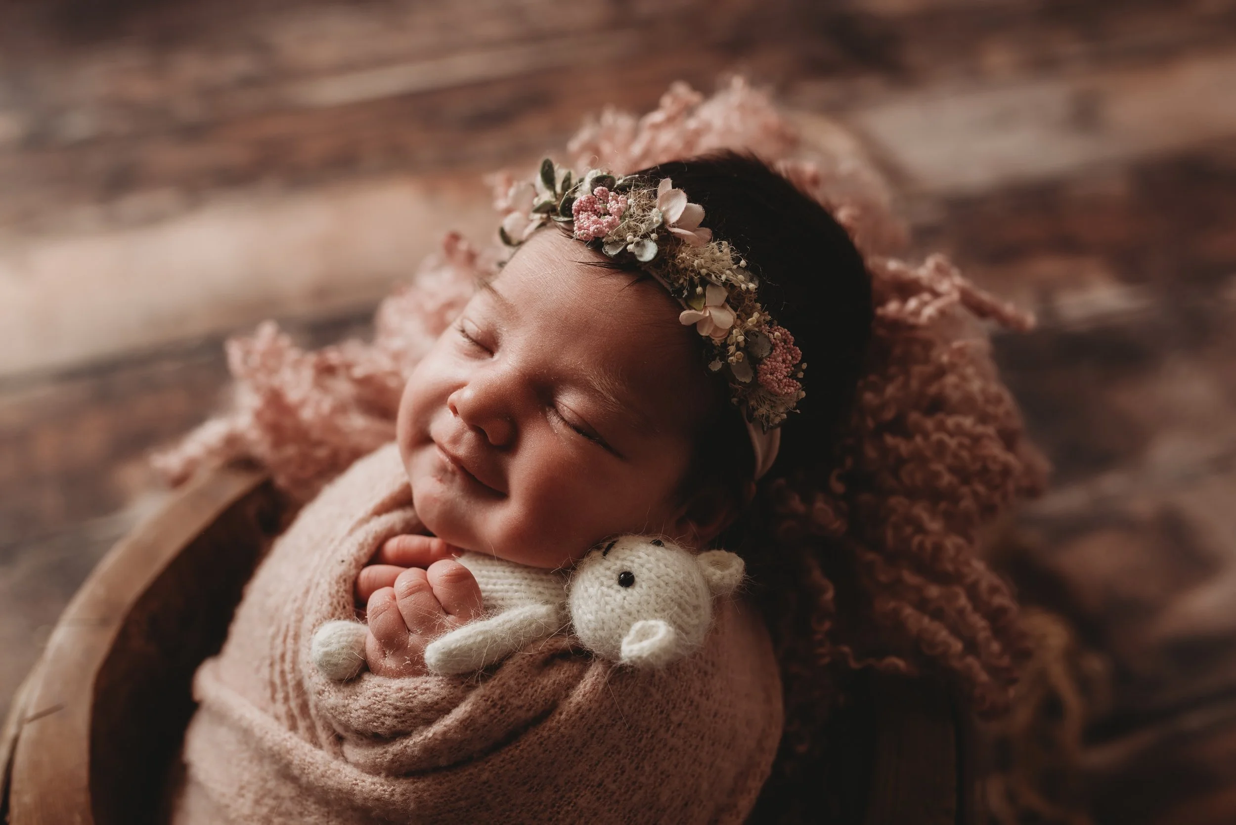 Newborn baby wrapped in pink blanket, wearing flower headband and holding knit bear.