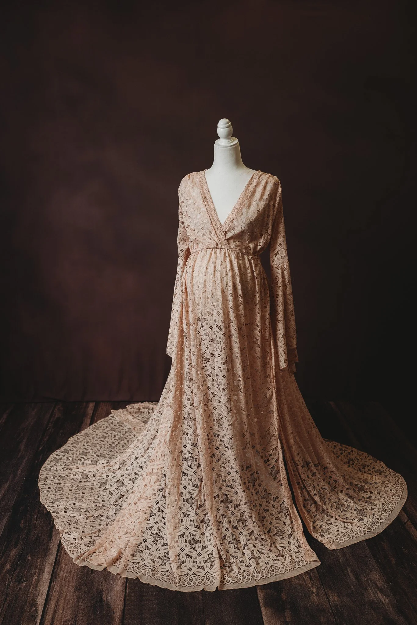 Mannequin wearing a long, lace, peach maternity gown on a wooden floor with a dark background.