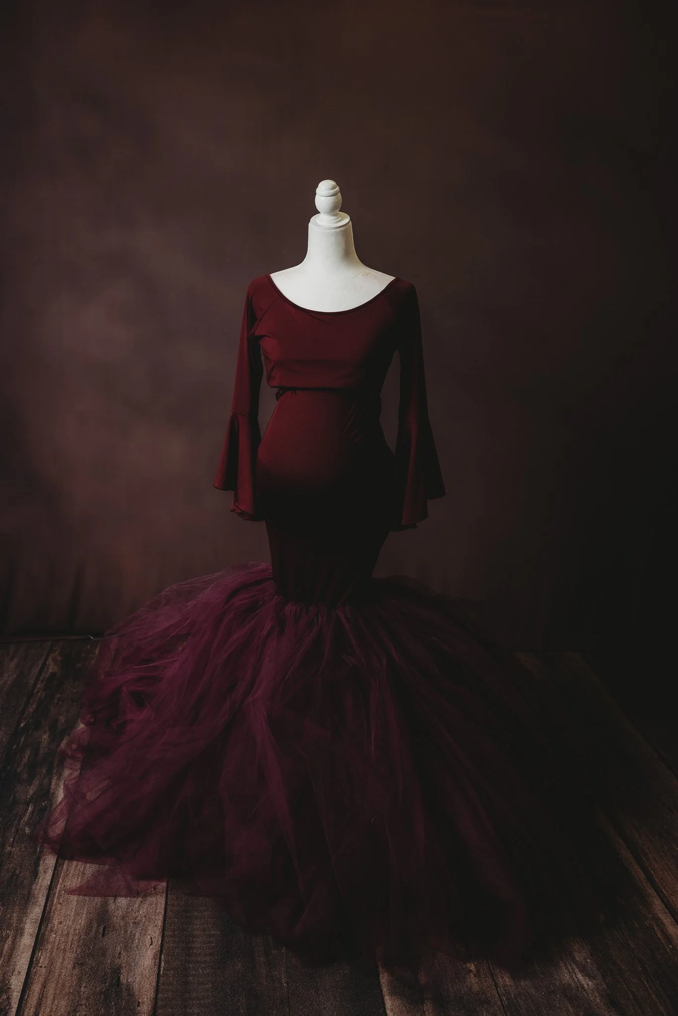 Mannequin wearing a burgundy mermaid maternity gown with flared sleeves on a wooden floor.
