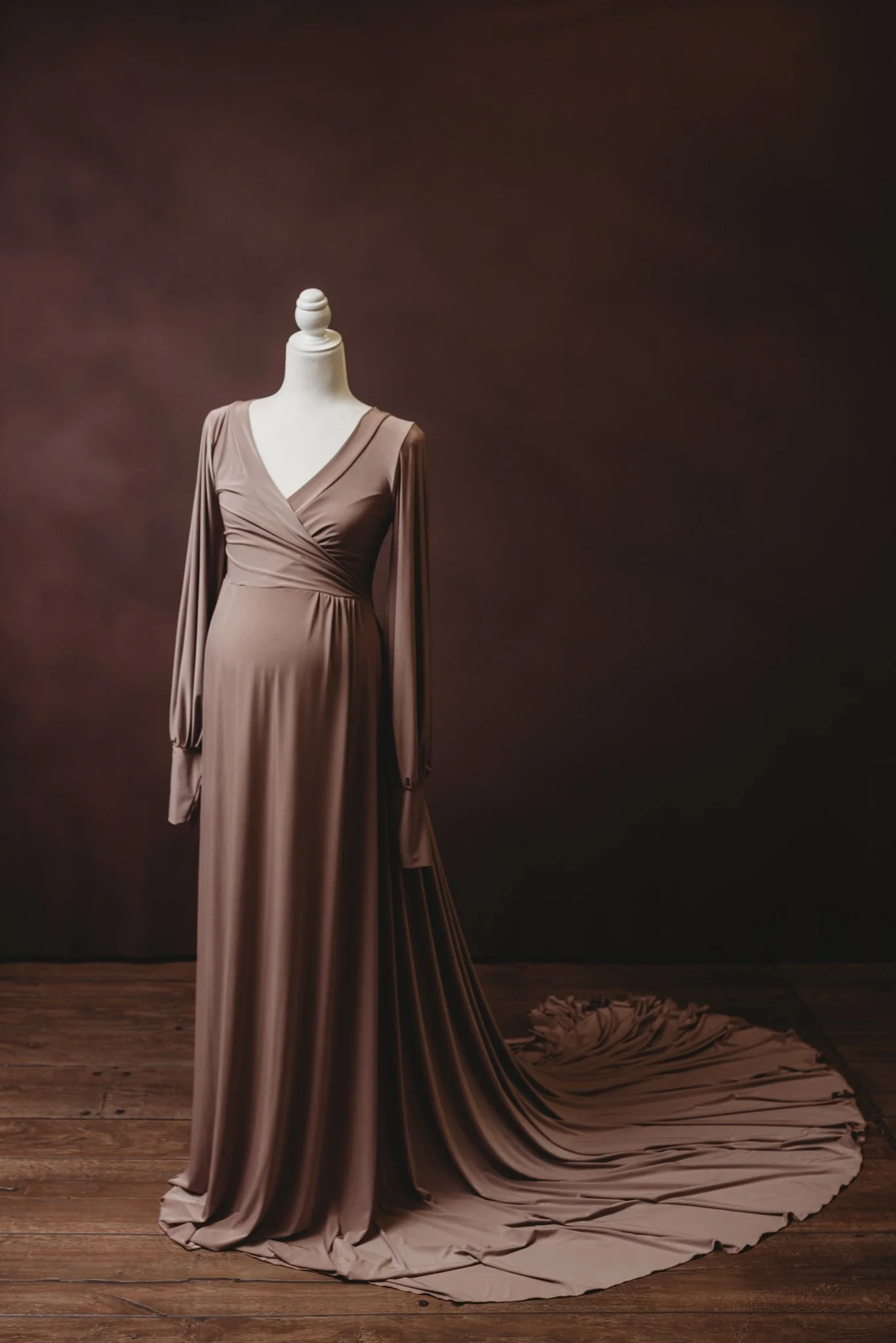 Elegant brown maternity gown with long sleeves and sweeping train on a mannequin, wooden floor backdrop.