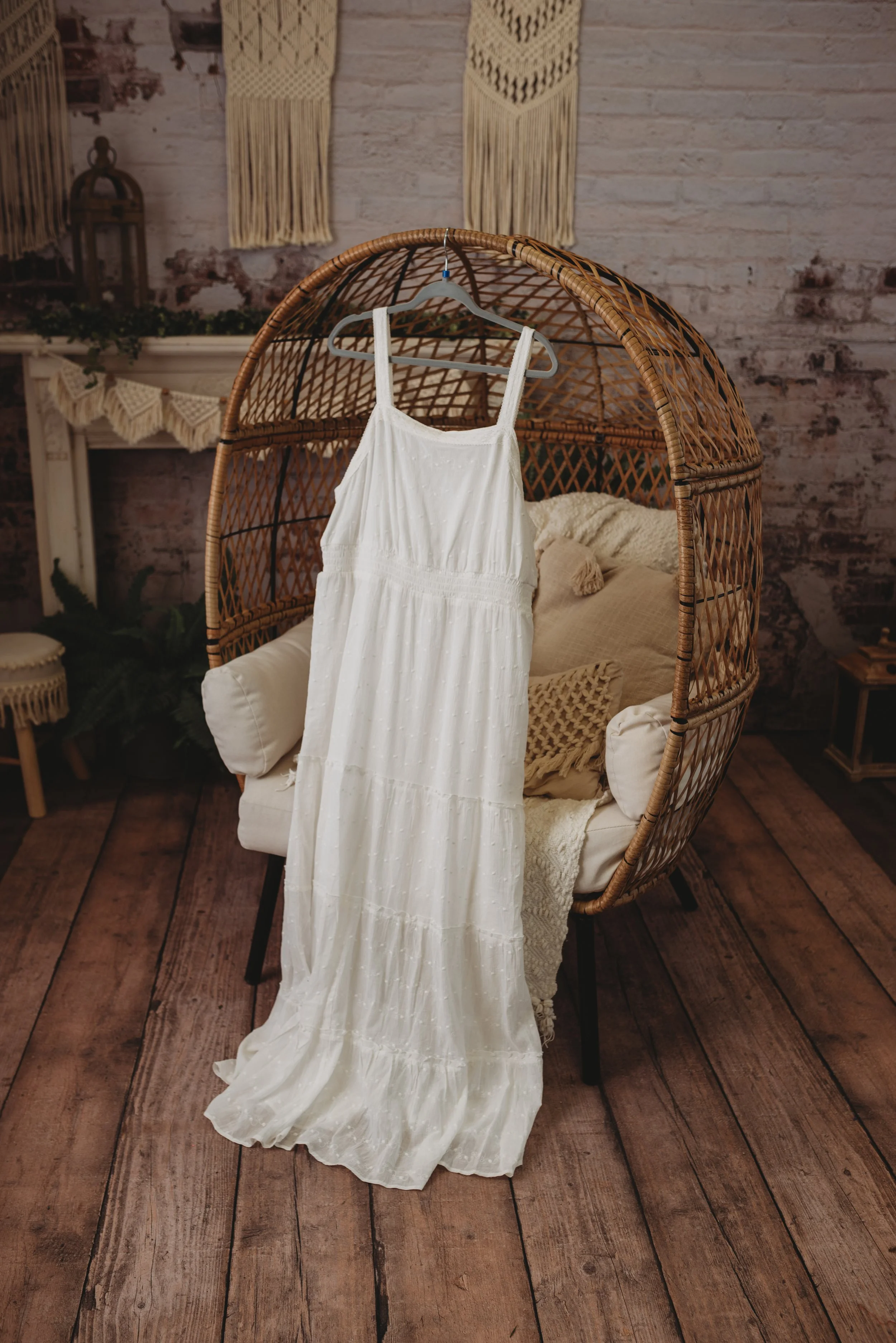 White dress hanging on a rattan chair in a rustic room with wooden flooring and macrame wall hangings.