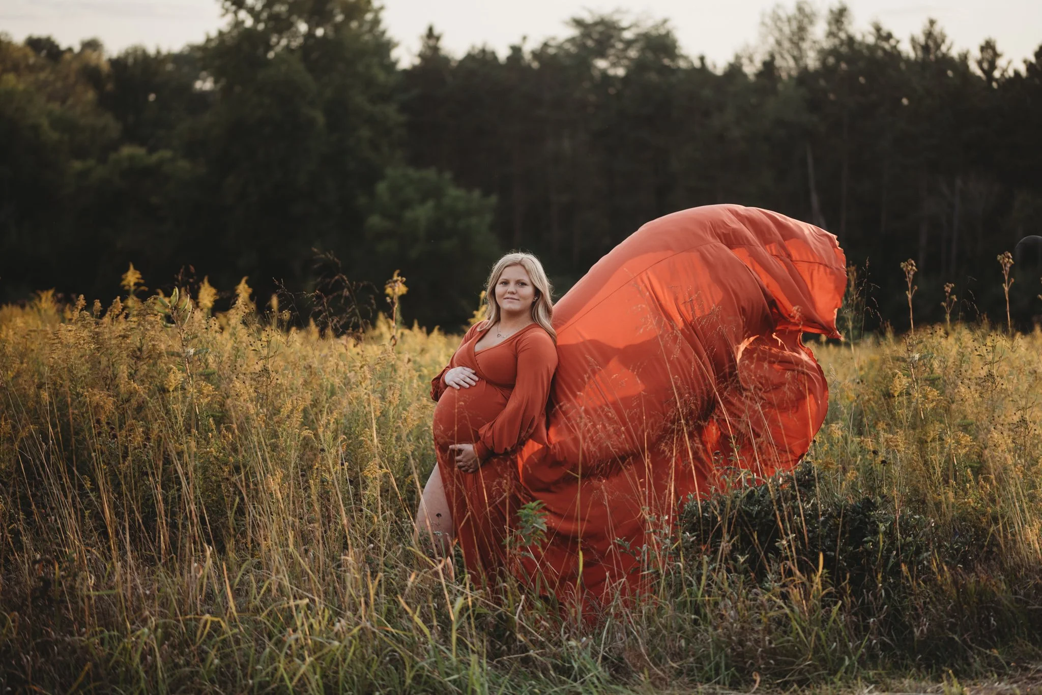 Pregnant woman in orange dress standing in field with flowing fabric, trees in background.