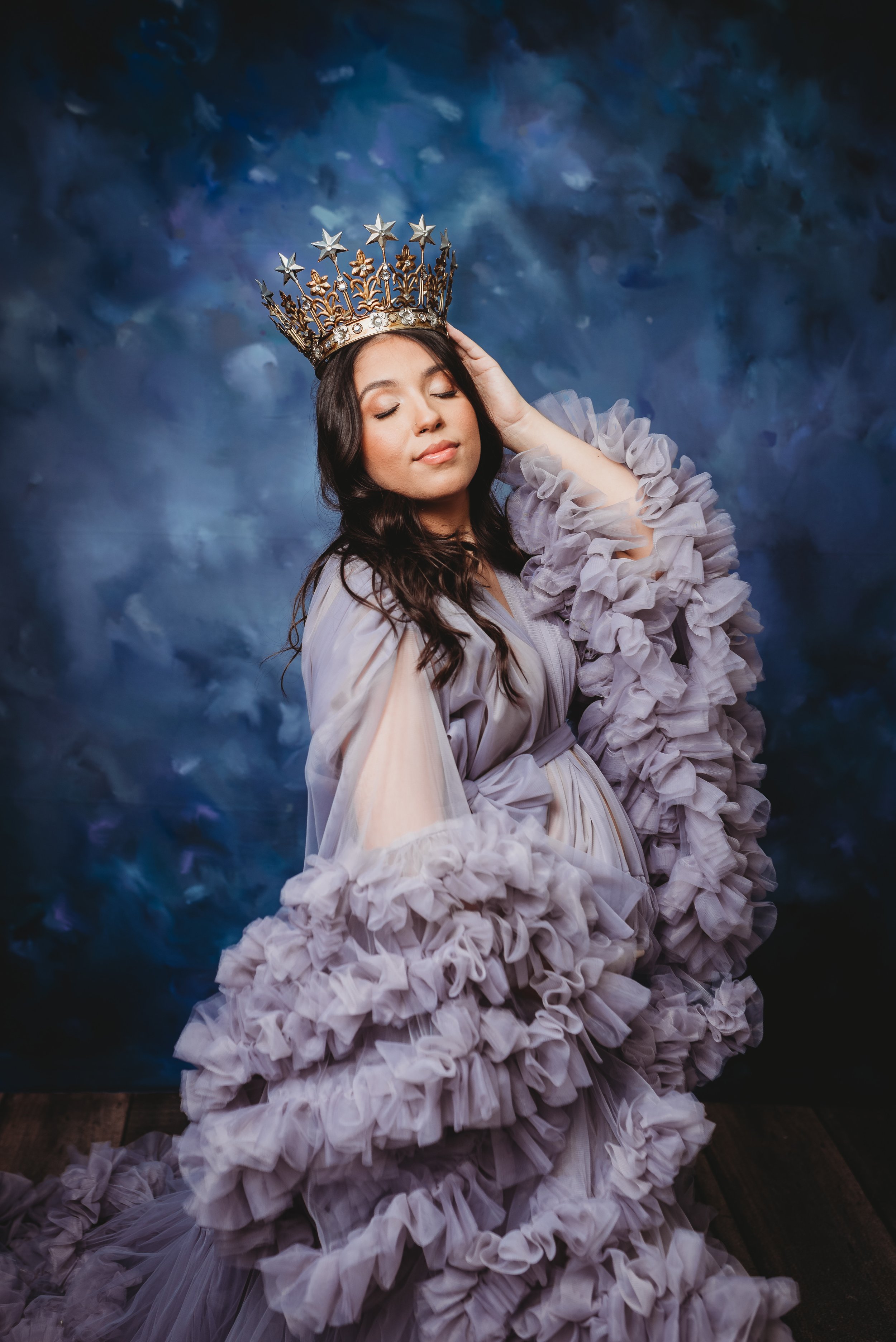 A woman with long, dark hair and closed eyes wearing a Gothic-style crown with silver stars and golden details. She is dressed in an elaborate gown with ruffled, layered sleeves, and a flowing, ruffled skirt. The background features a dark, cloudy, b