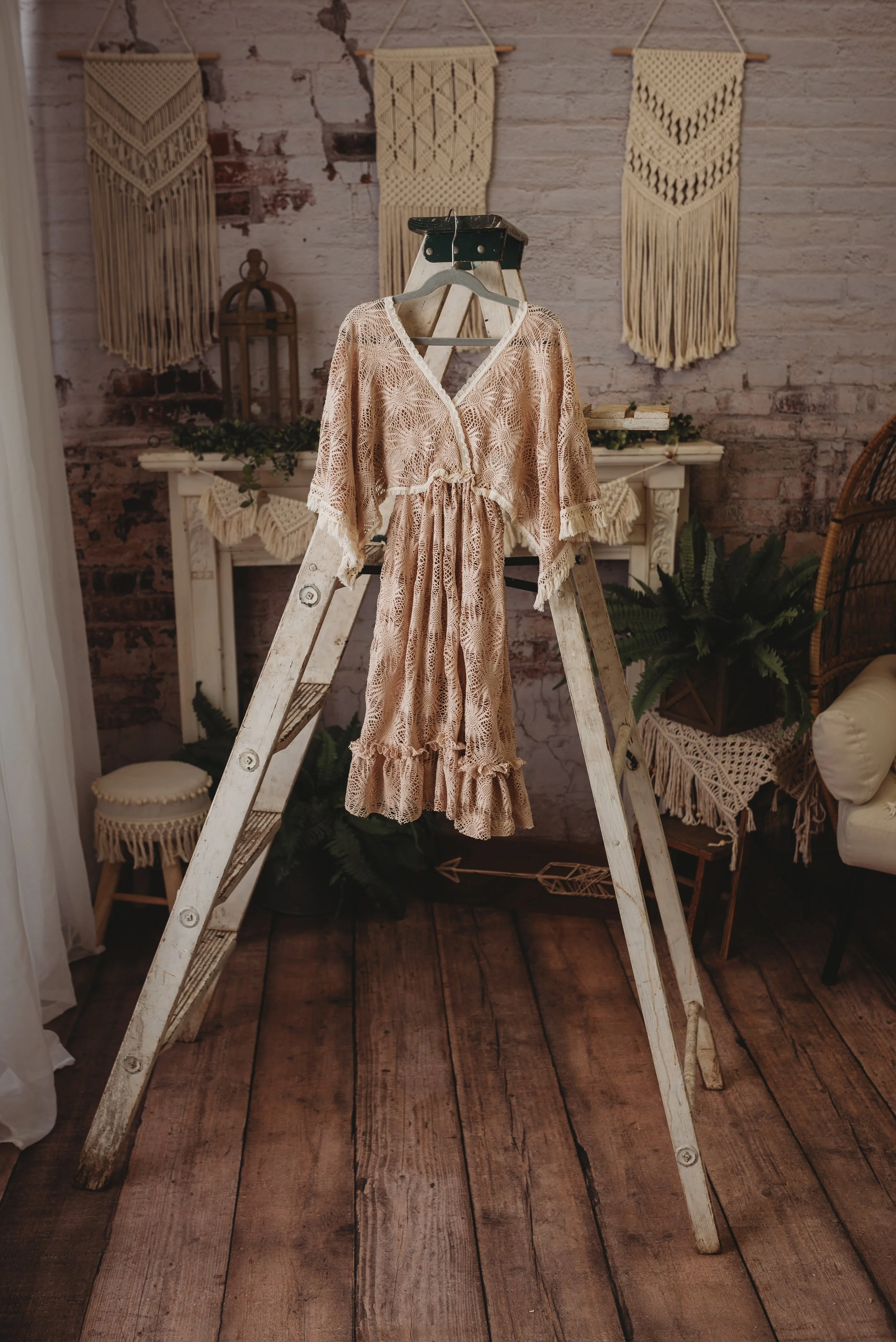 Bohemian beige lace dress hung on a rustic wooden ladder in a vintage room with macramé wall hangings and plants.