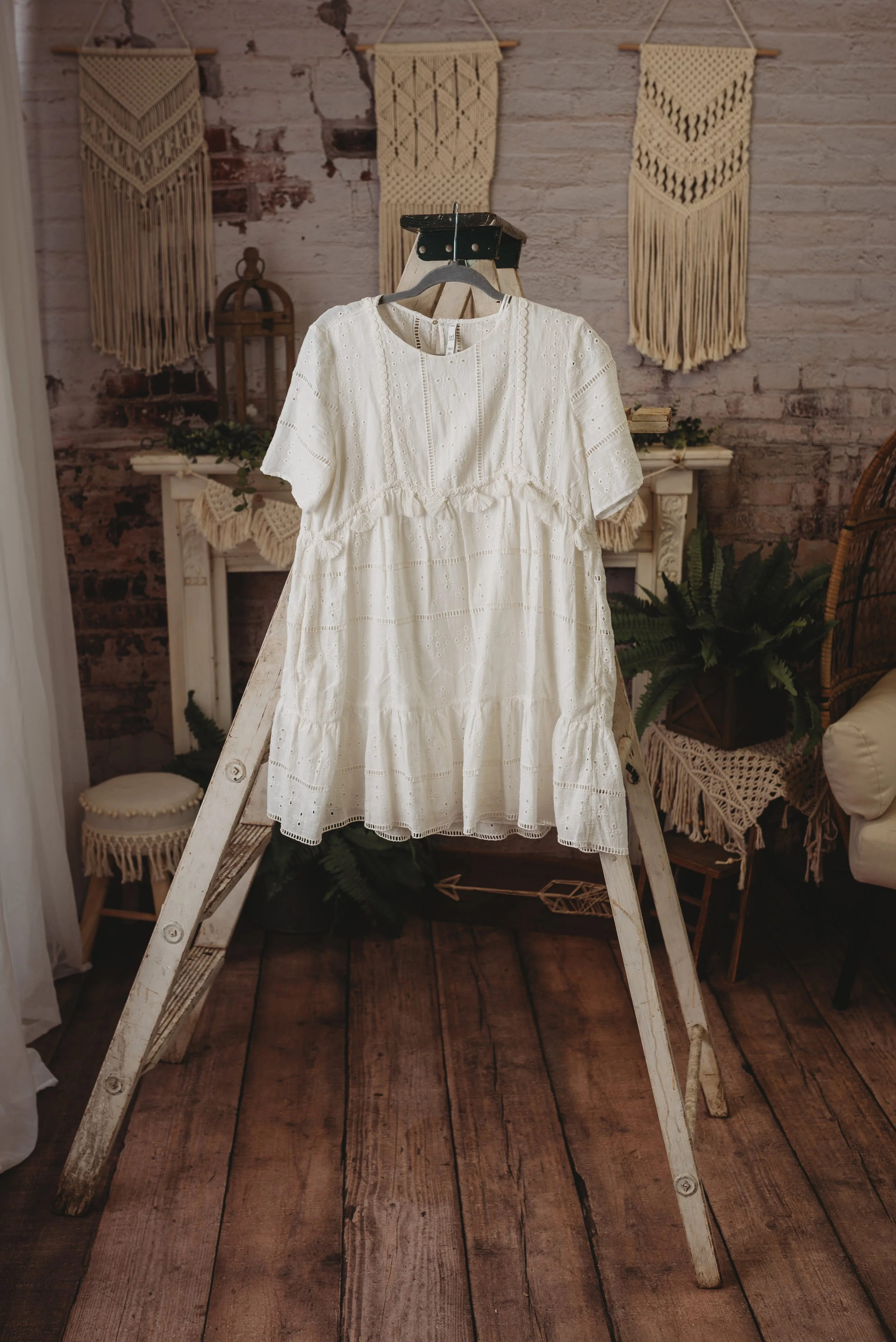 White lace dress on hanger with vintage decor background, including wooden ladder, potted plants, and macramé wall hangings.