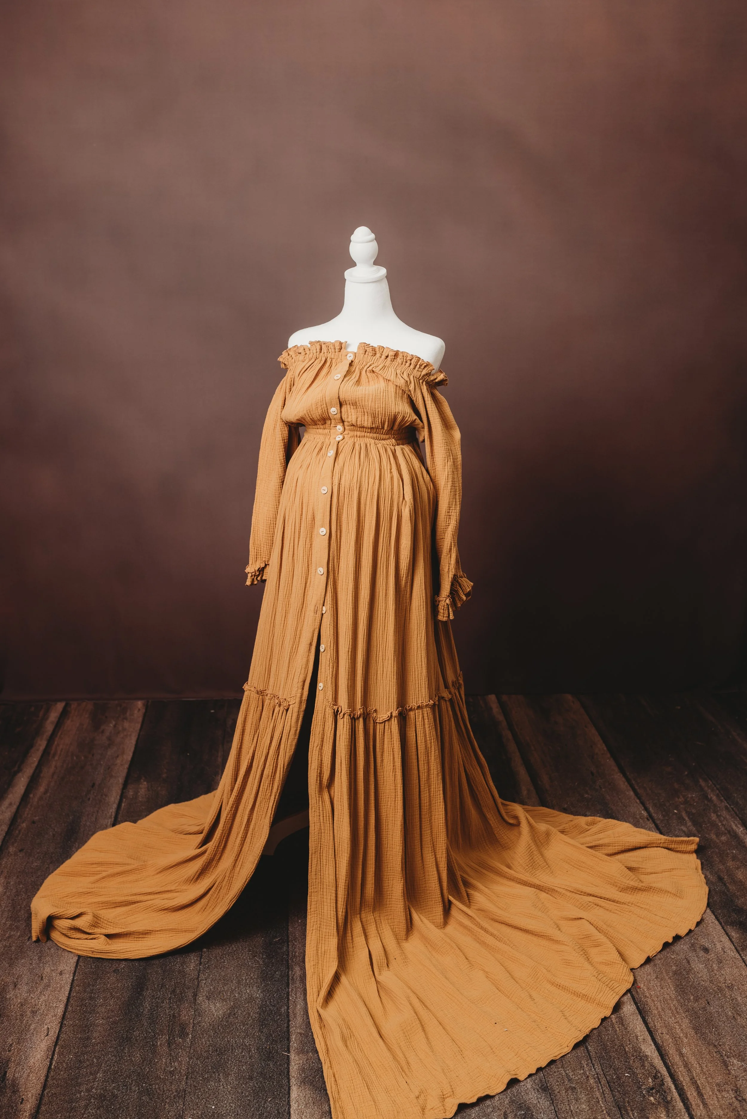 A beige, off-shoulder, button-down dress with ruffled details on a dress form mannequin, with long flowing fabric trailing on a dark wooden floor, against a brown backdrop.