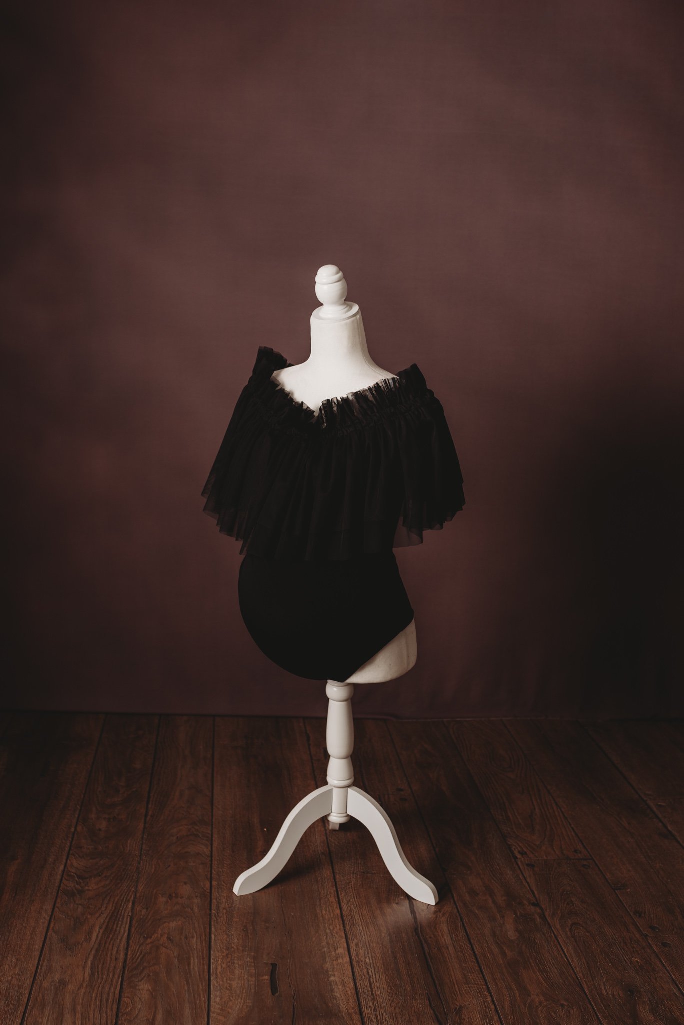 Vintage white dress form mannequin with black ruffled fabric on a wooden floor and brown background.