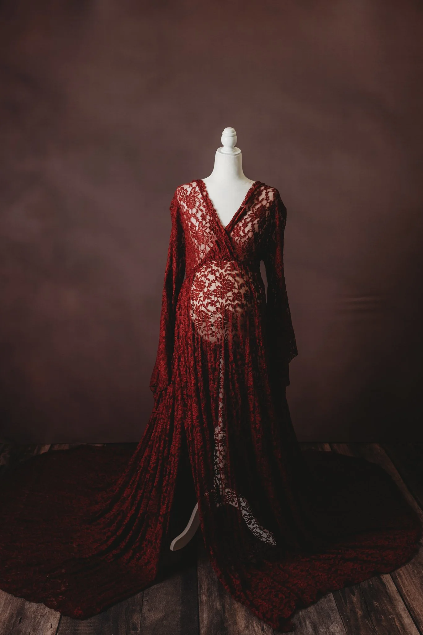 Mannequin wearing a burgundy lace maternity gown with floral pattern, long sleeves, and a flowing train, set against a dark backdrop.