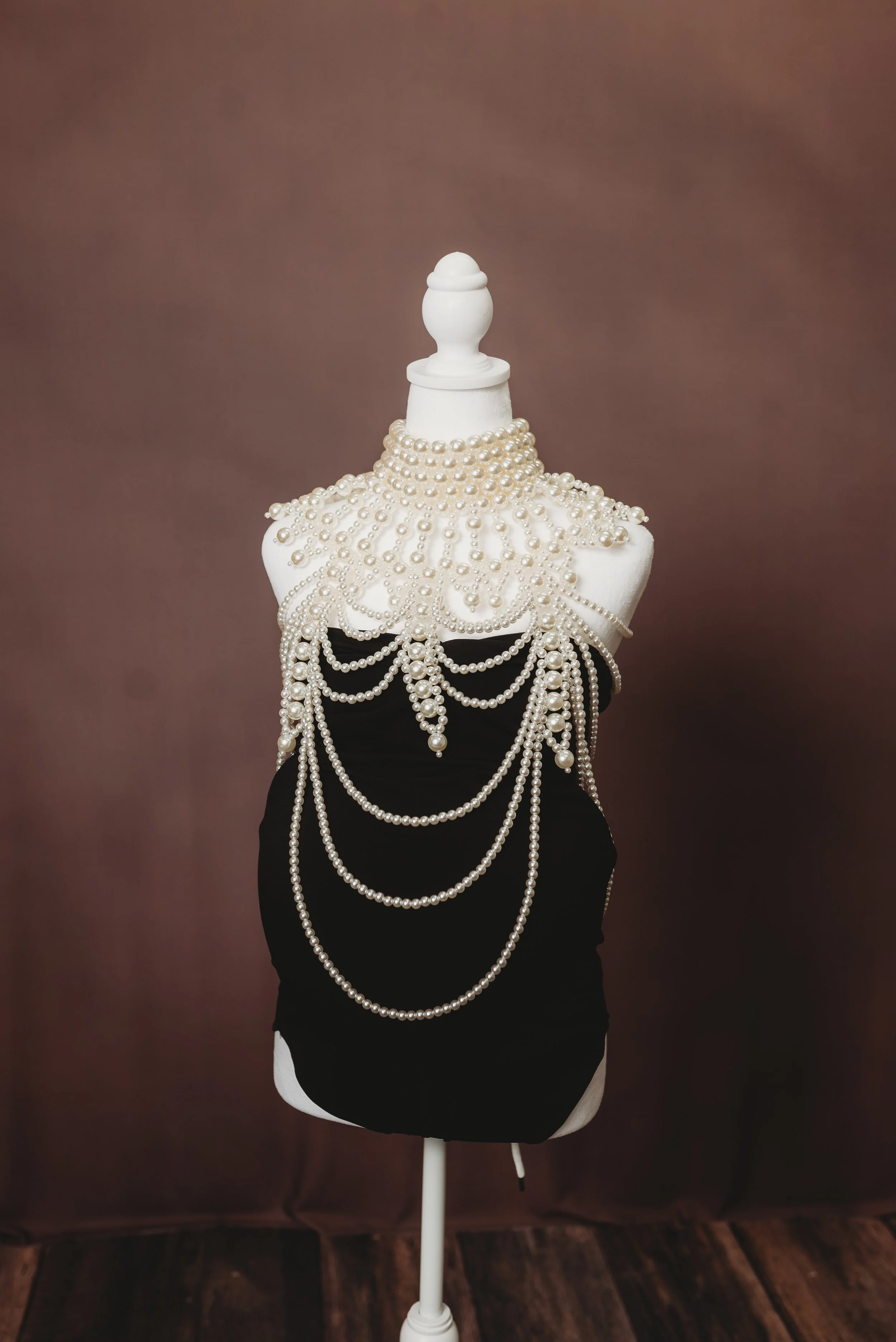 White mannequin torso adorned with multiple strands of pearl necklaces, draped over a black fabric. The mannequin has a white decorative finial at the top and is set against a brown background with a wooden floor.