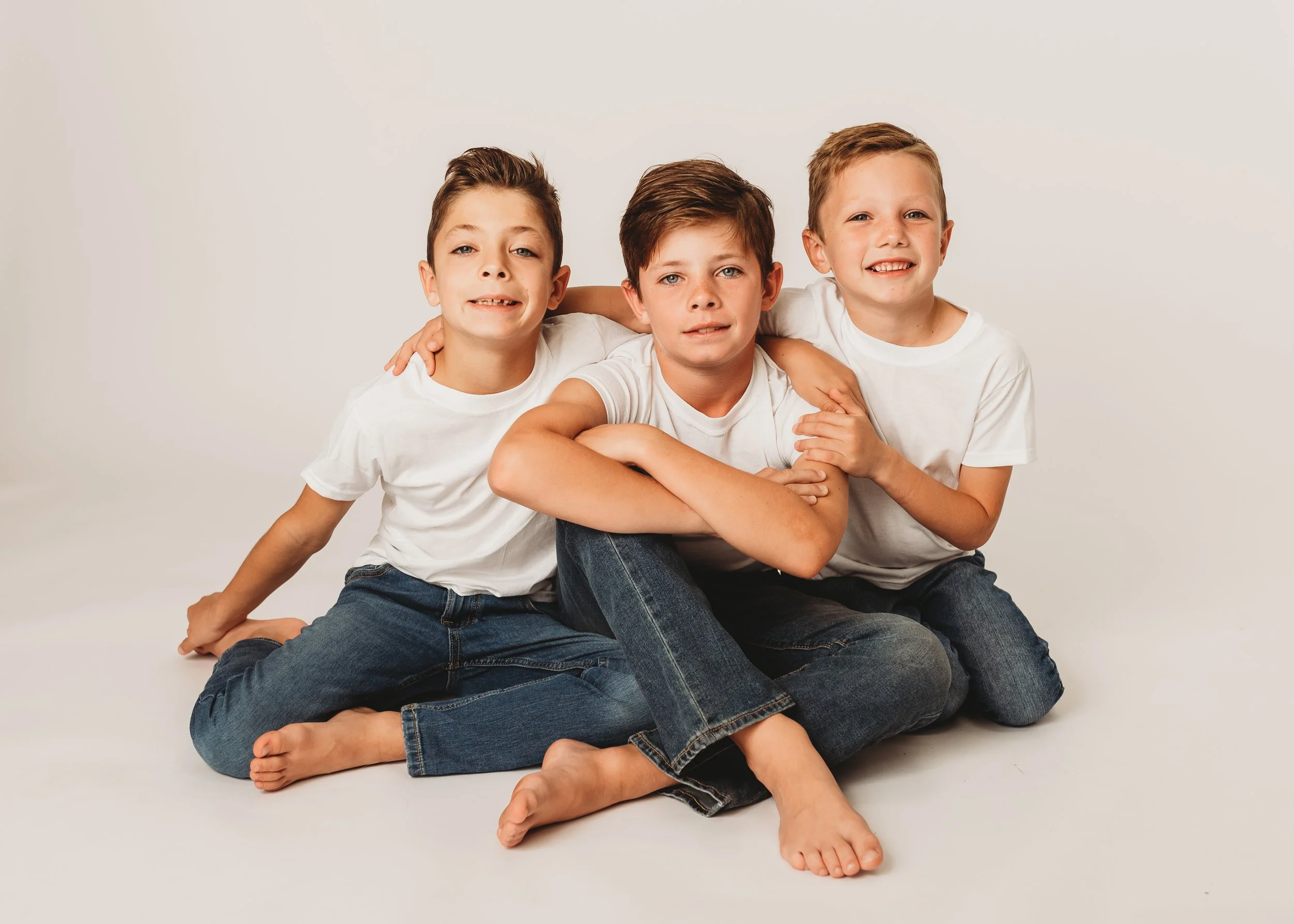 Three boys sitting on floor in white shirts and jeans, smiling.