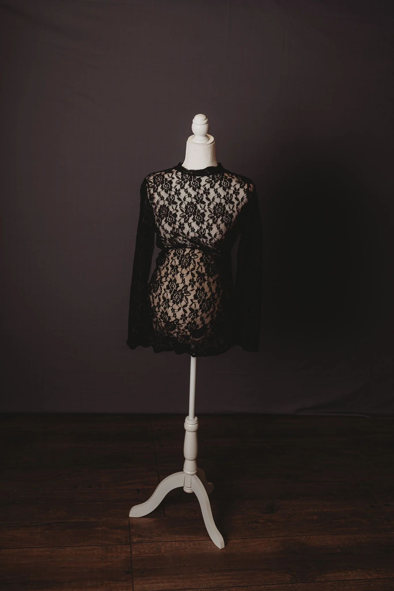 White mannequin wearing a black lace maternity dress with floral patterns, displayed on a wooden stand against a dark background.