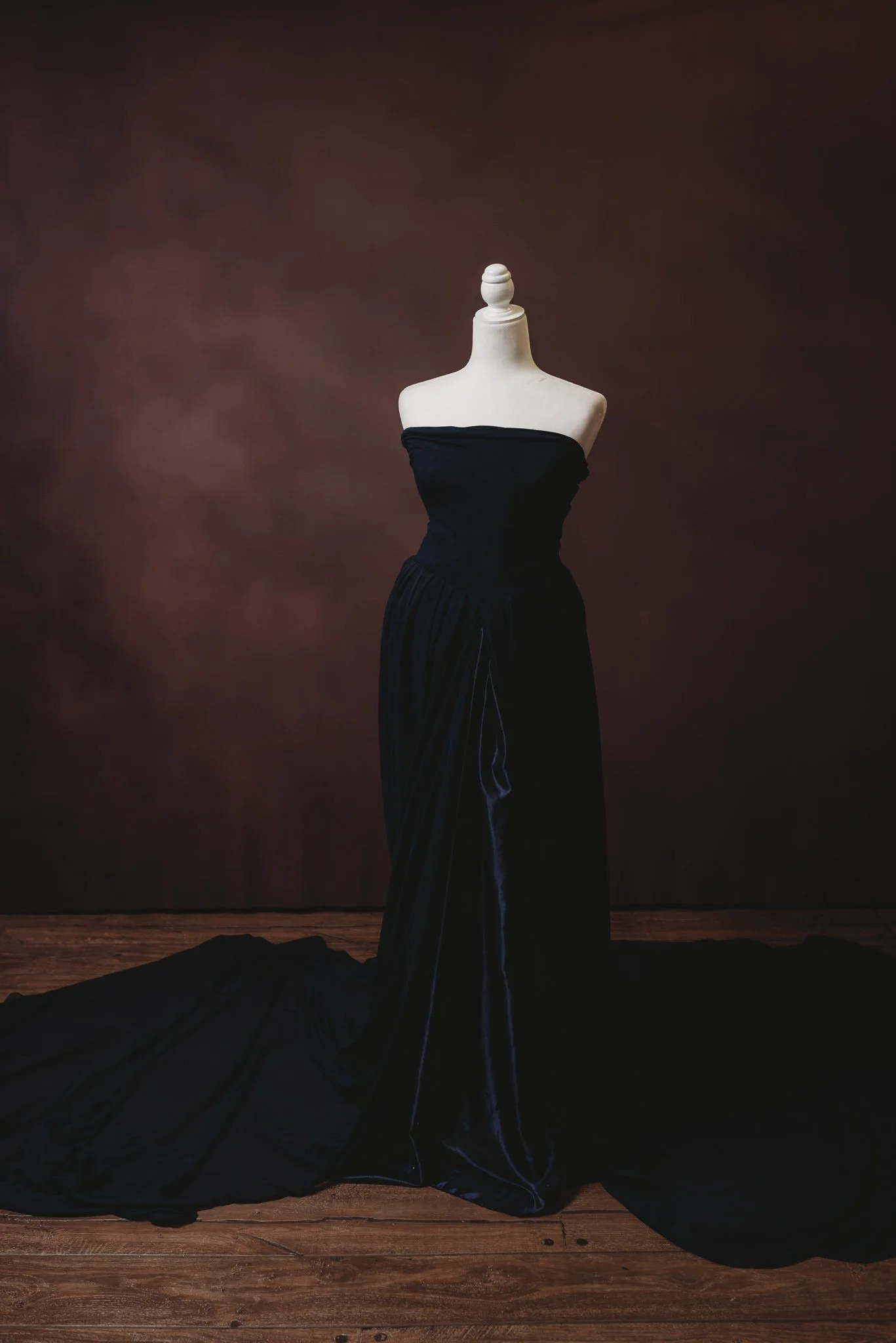 Mannequin displaying a strapless black velvet evening gown with a long train on a wooden floor and a dark brown background.
