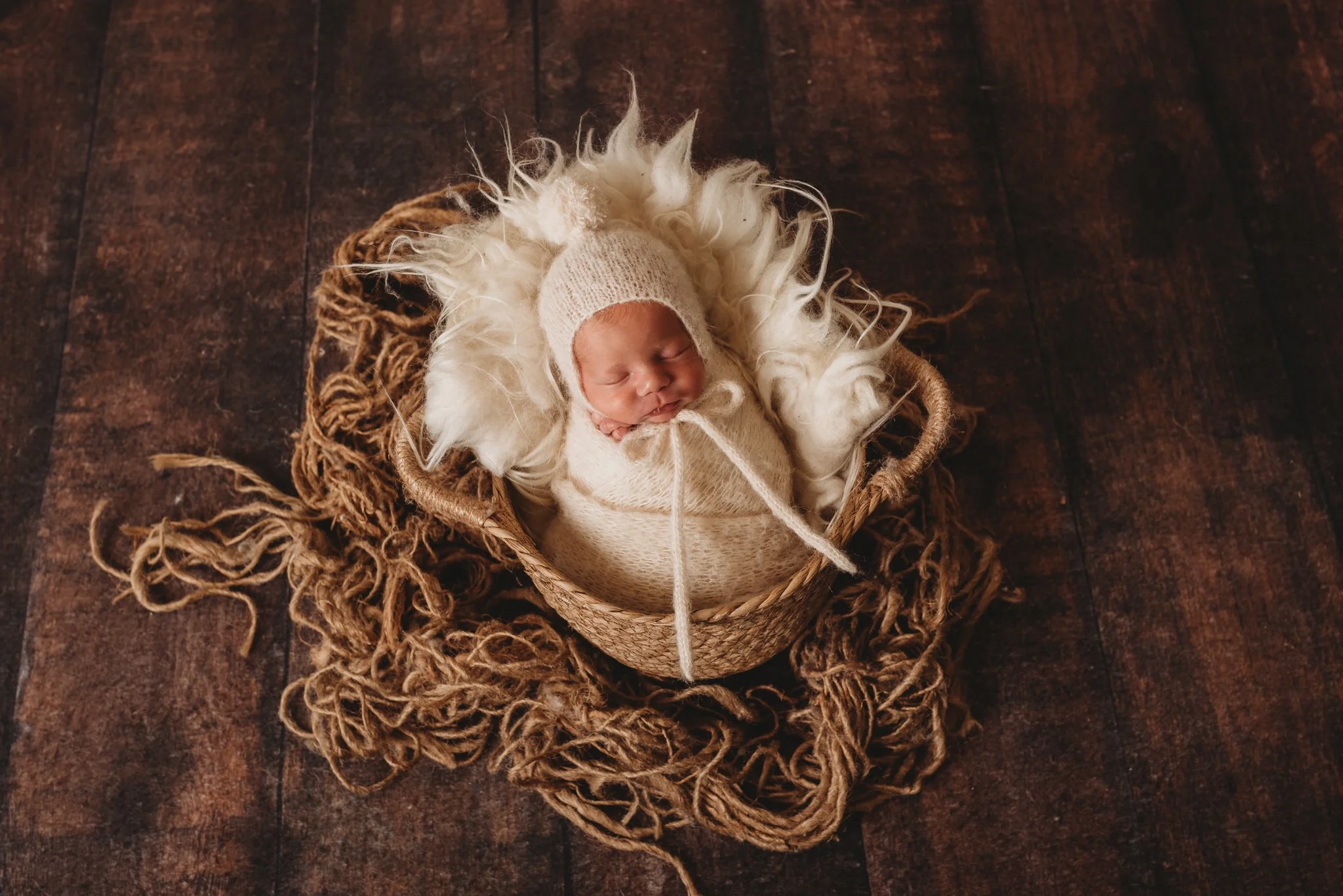 Newborn baby sleeping in a basket, wrapped in a cream-colored blanket with knitted hat, surrounded by fluffy material and rustic rope accents.