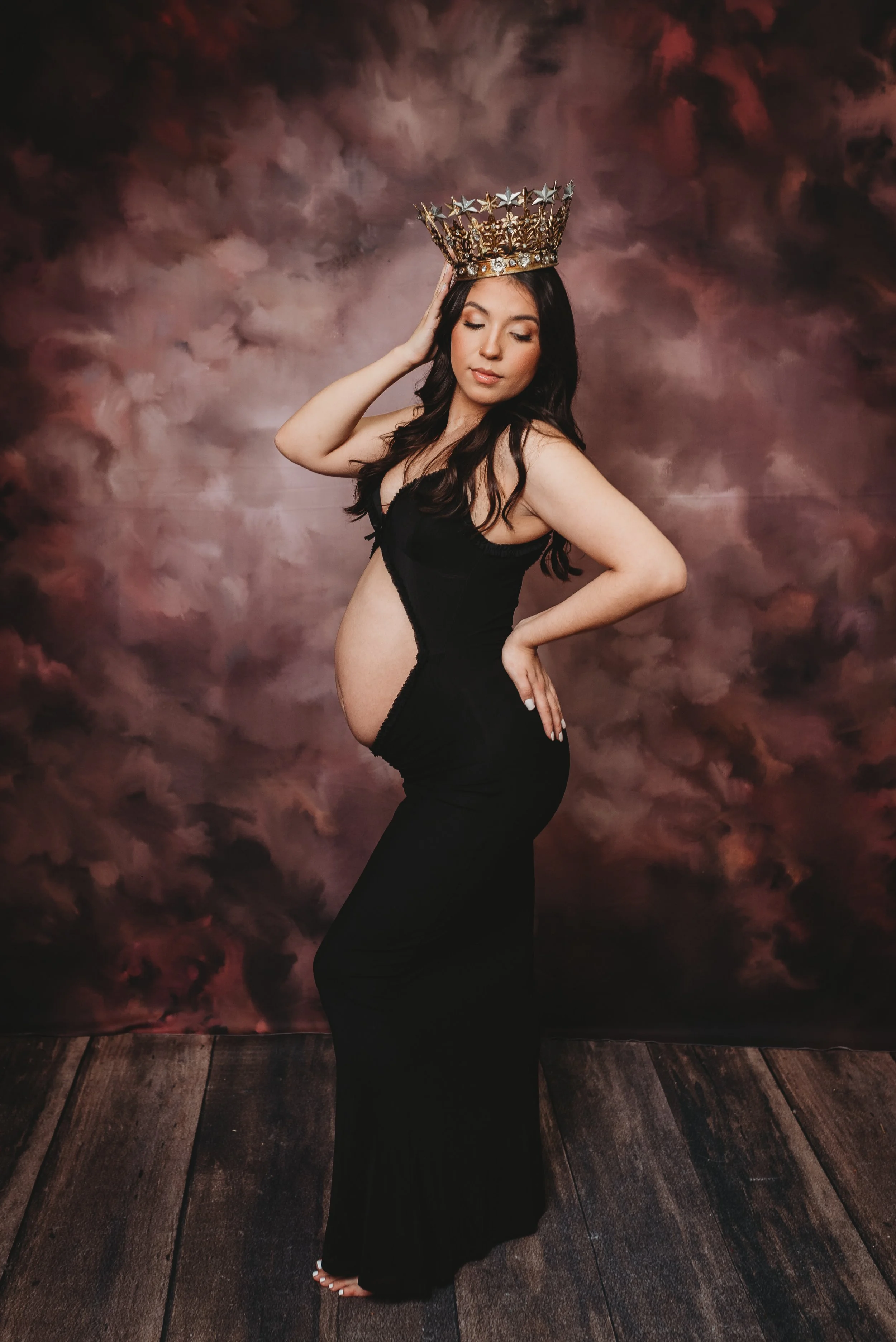 A pregnant woman in a black dress with a cutout, standing barefoot on a wooden floor, wearing an ornate crown, with her eyes closed and one hand on her hip, posed in front of a cloudy pink and purple background.