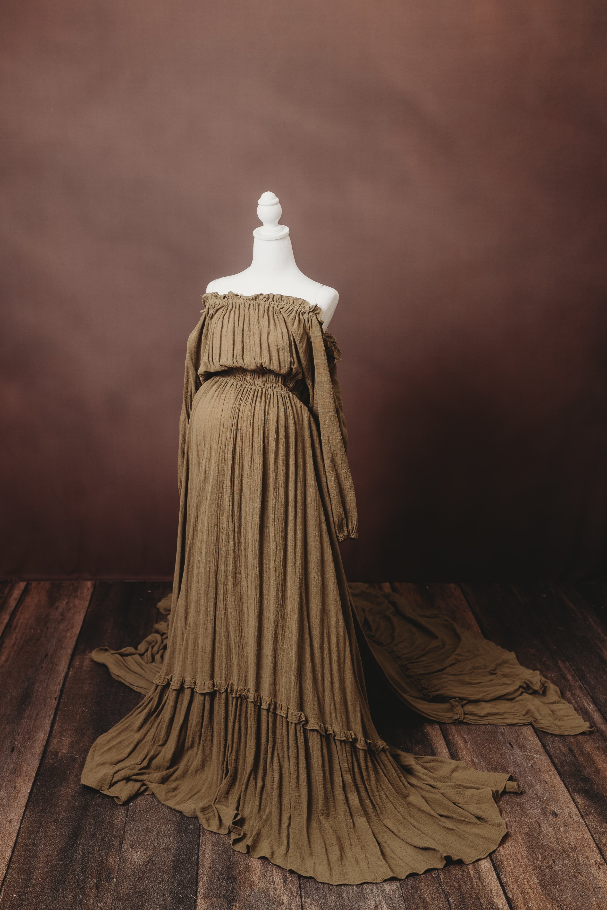 A white dress form mannequin draped with a brown pleated, off-the-shoulder gown, standing on a wooden floor with a plain brown background.