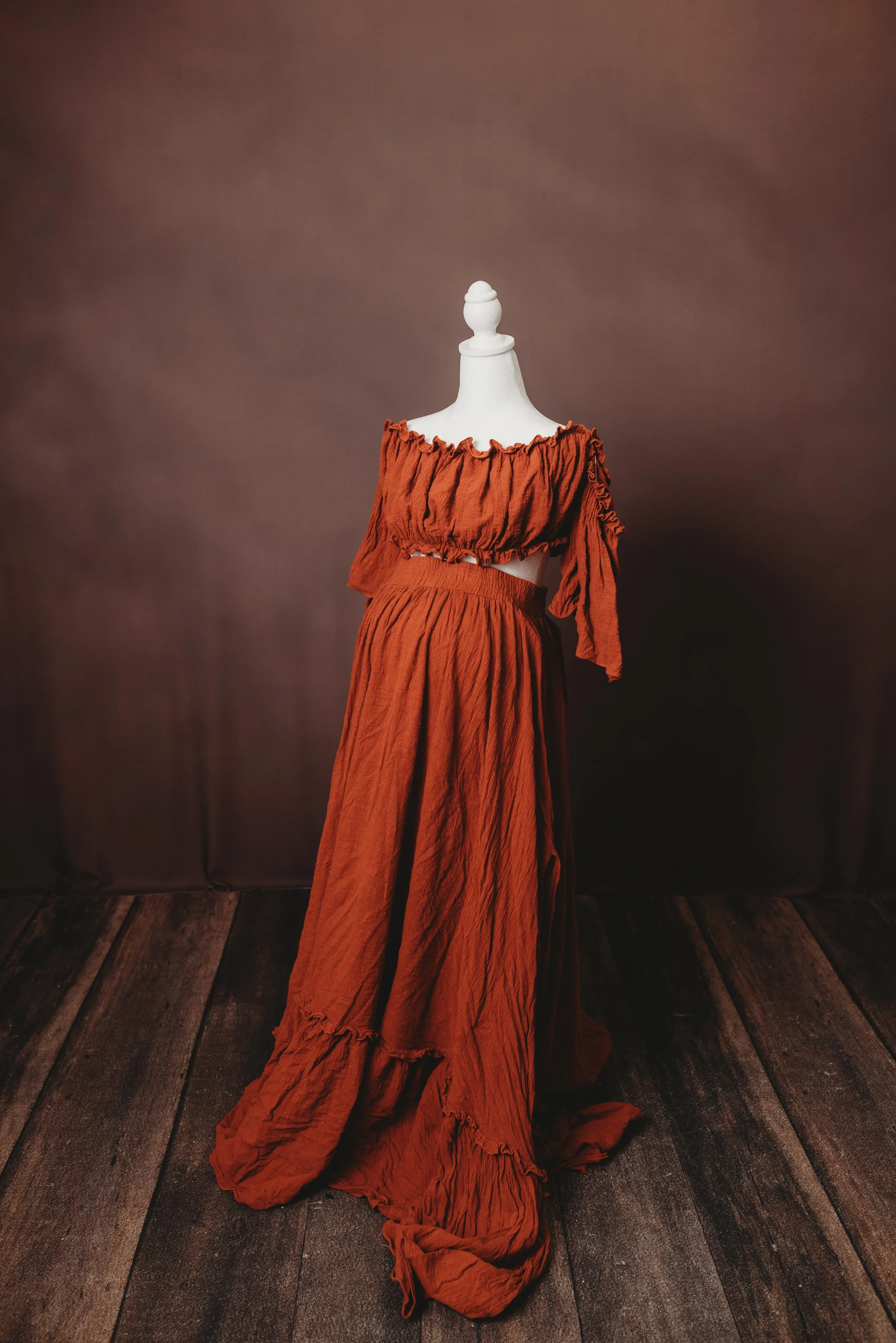 A mannequin dressed in an off-the-shoulder, burnt orange, crinkled fabric maxi dress with ruffled edges, displayed in front of a brown backdrop on a wooden floor.