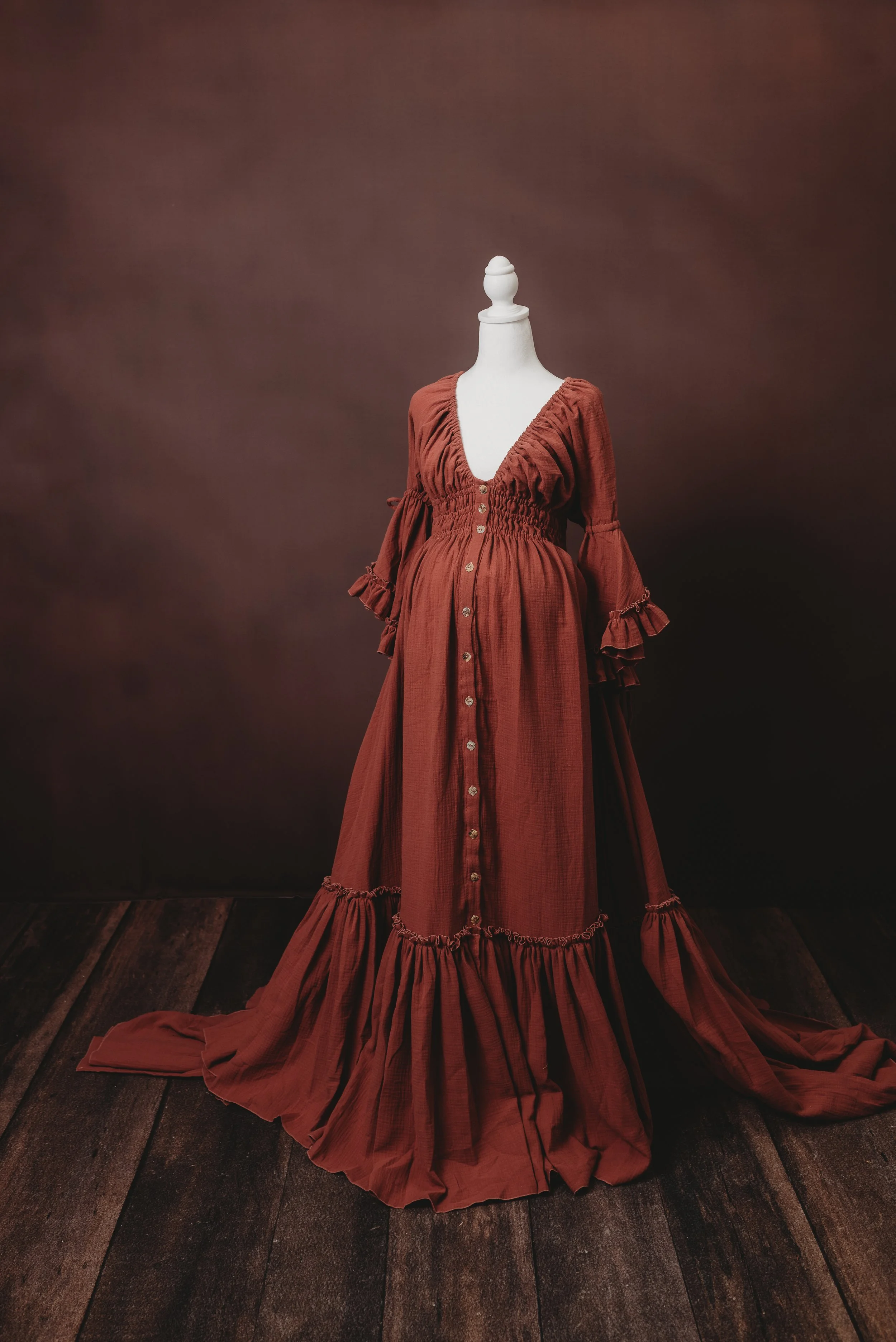 A mannequin dressed in a long, rust-colored vintage-style dress with ruffled sleeves and a button-down front, photographed against a brown background and standing on a wooden floor.