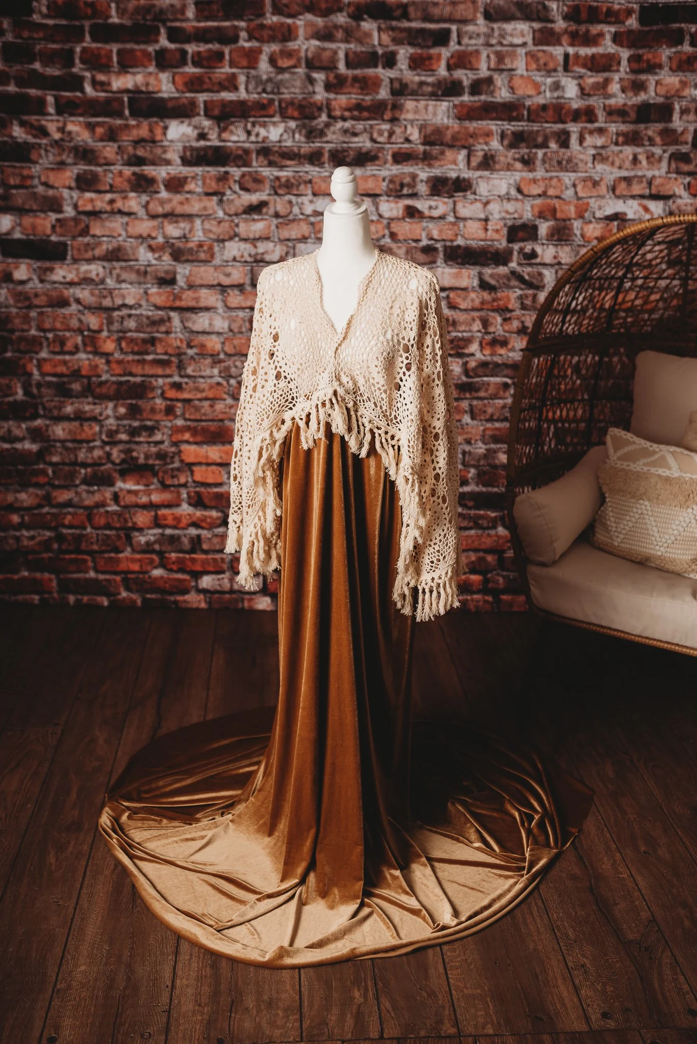 Mannequin displaying a beige crocheted shawl and long gold velvet skirt, set against a brick wall background with a wicker chair.