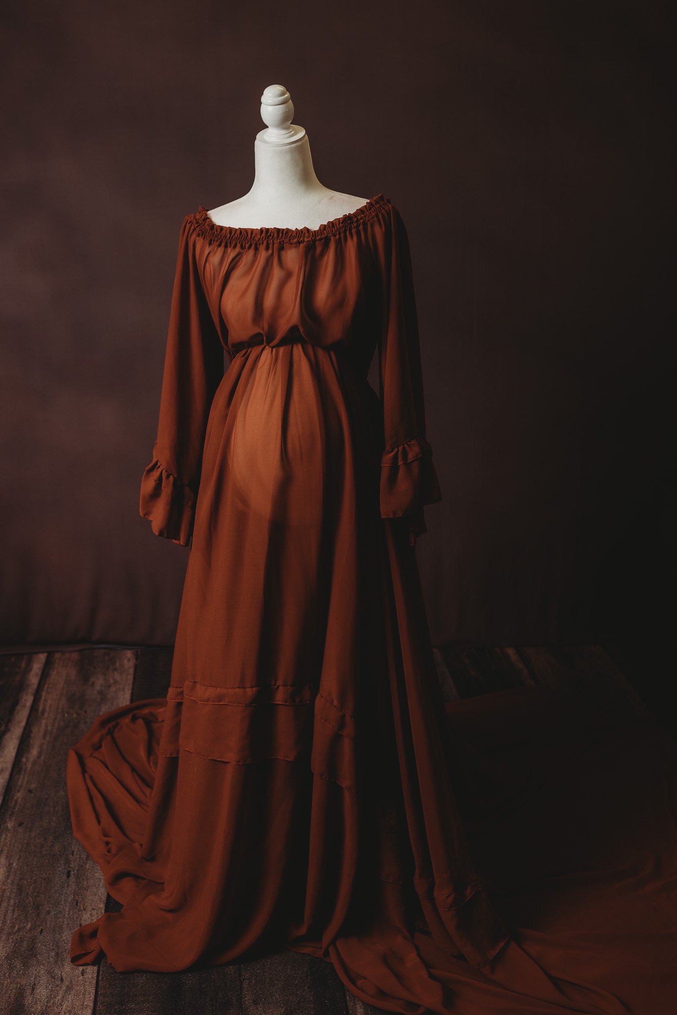 Mannequin wearing a long, off-shoulder brown dress with ruffled sleeves and train, displayed against a dark backdrop.